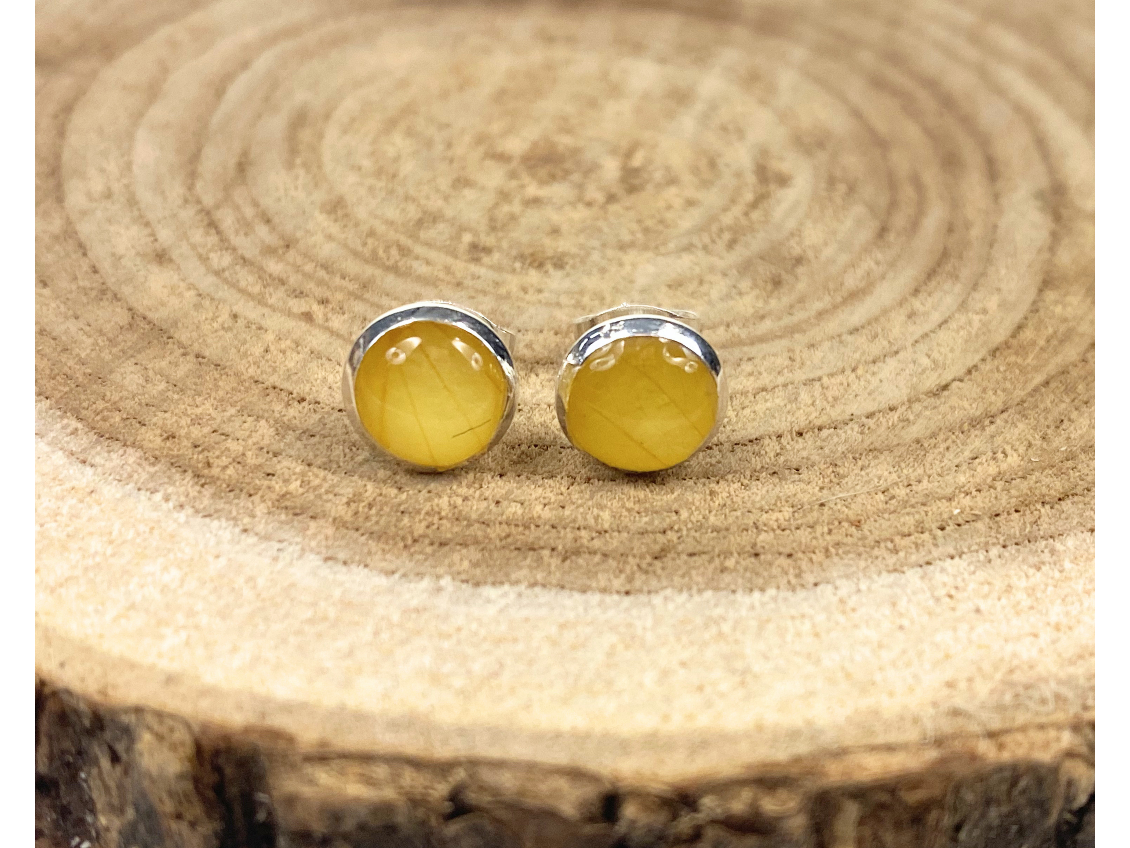 A pair of round silver stud earrings containing yellow flower petals, displayed on a rustic wood slice background. 
