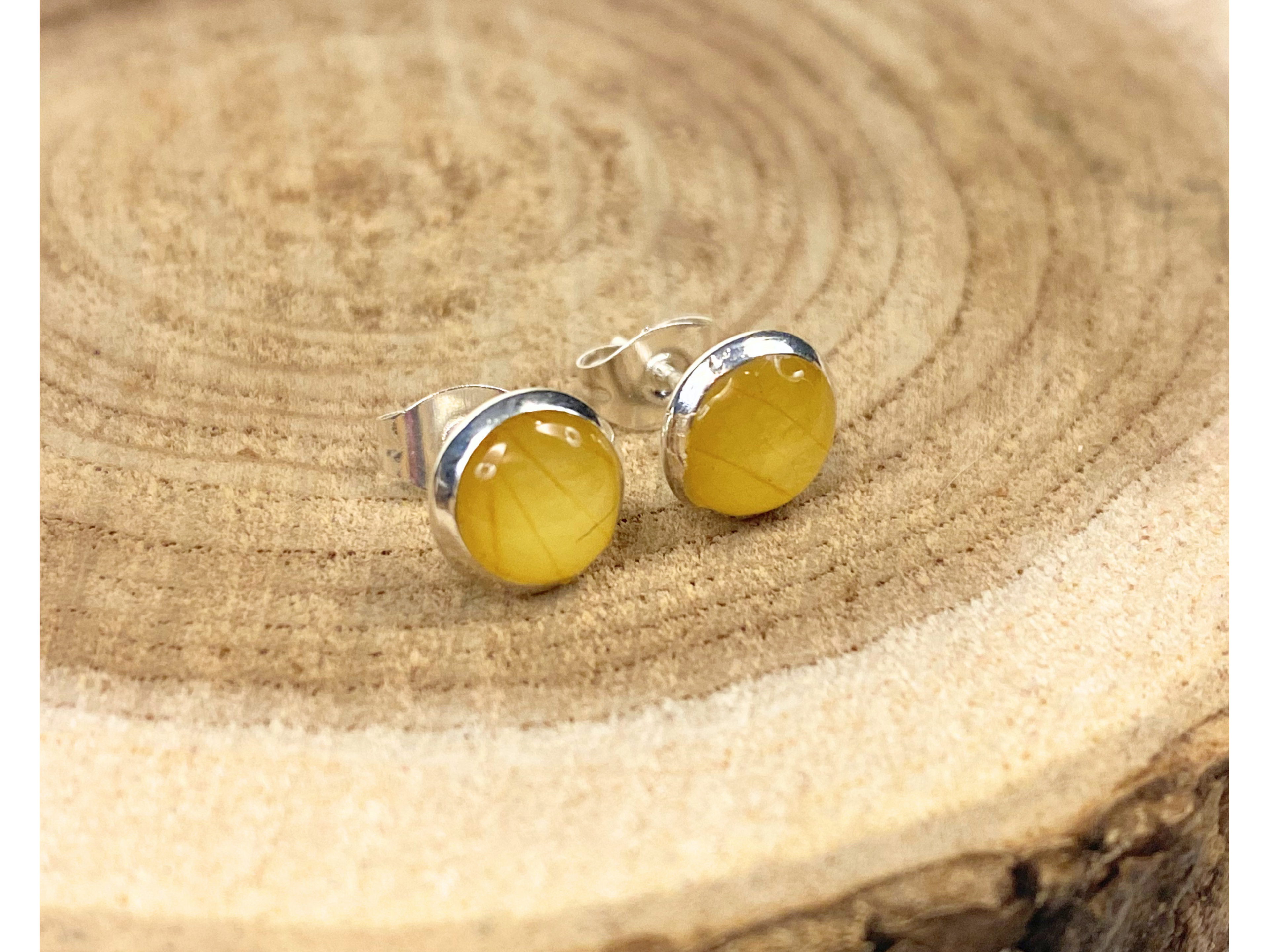 A pair of round silver stud earrings containing yellow flower petals, displayed on a rustic wood slice background. 