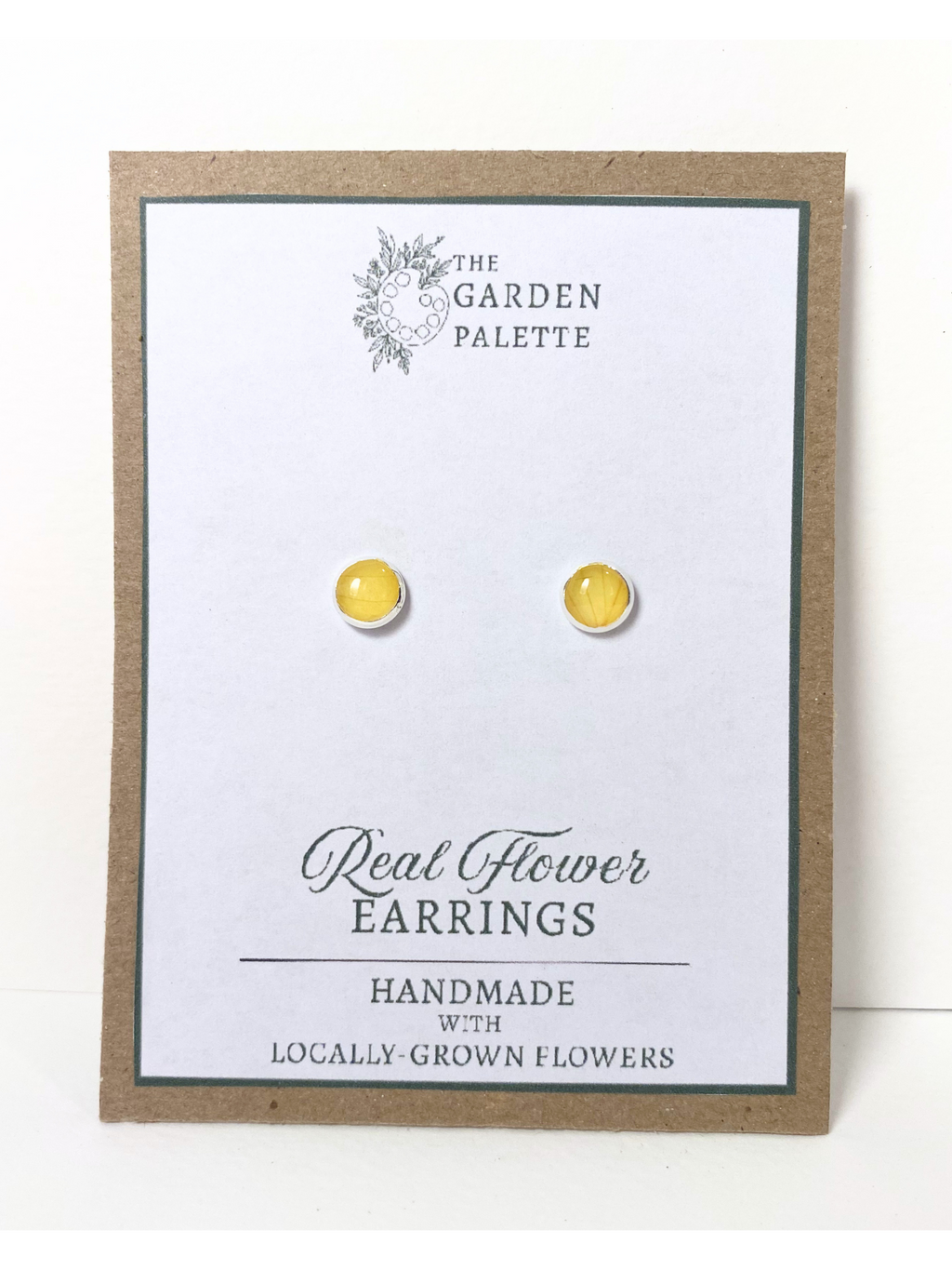 A pair of round silver stud earrings containing yellow flower petals, displayed on a white card mounted on a kraft paper backing. The card reads 'The Garden Palette' and 'Real Flower Earrings' with 'Handmade with Locally Grown Flowers.'