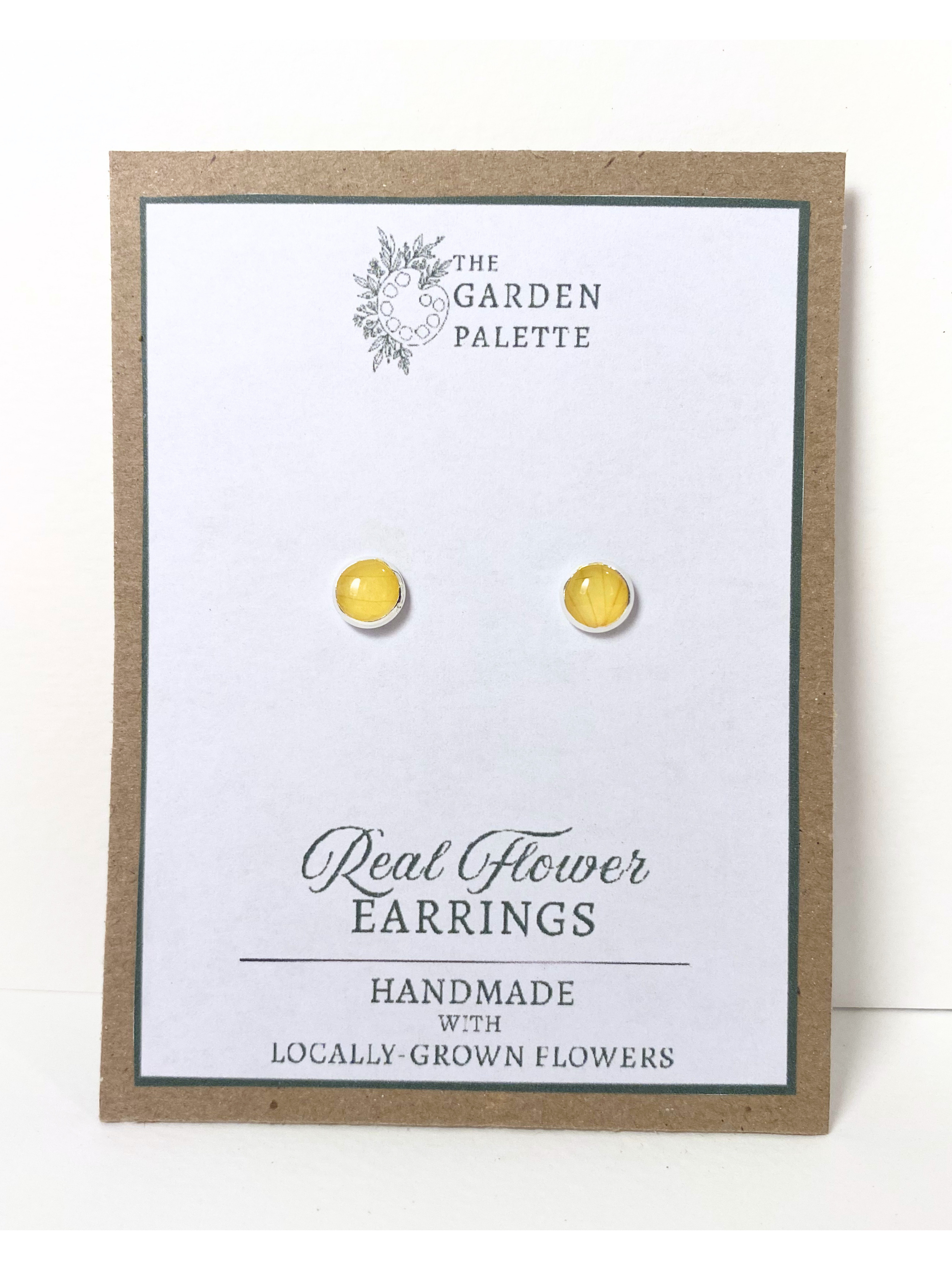 A pair of round silver stud earrings containing yellow flower petals, displayed on a white card mounted on a kraft paper backing. The card reads 'The Garden Palette' and 'Real Flower Earrings' with 'Handmade with Locally Grown Flowers.'
