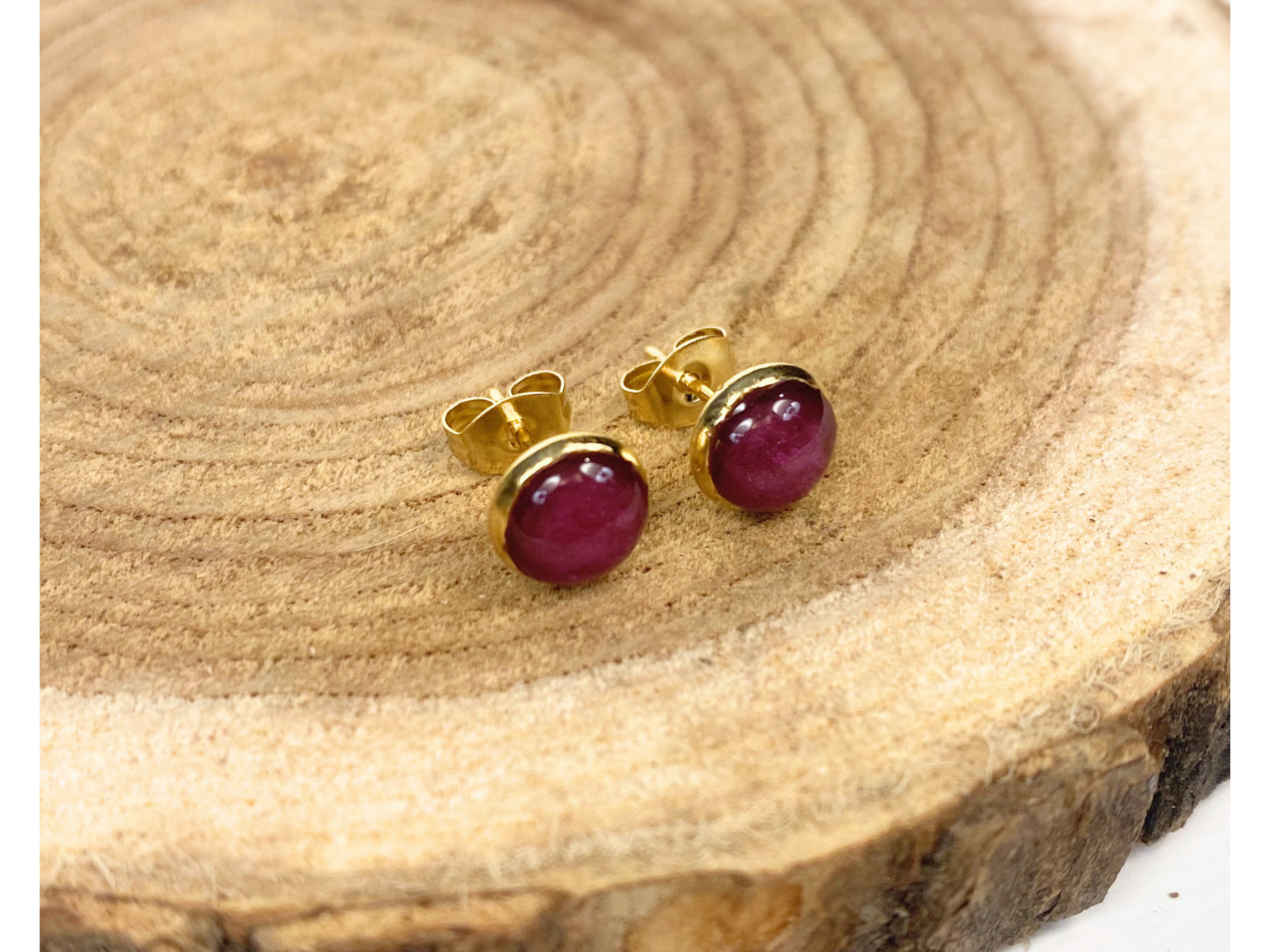 A pair of gold stud earrings containing purple flower petals, displayed on a rustic wood slice background.