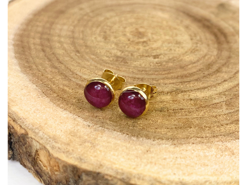 A pair of gold stud earrings containing purple flower petals, displayed on a rustic wood slice background.