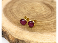 A pair of gold stud earrings containing purple flower petals, displayed on a rustic wood slice background.