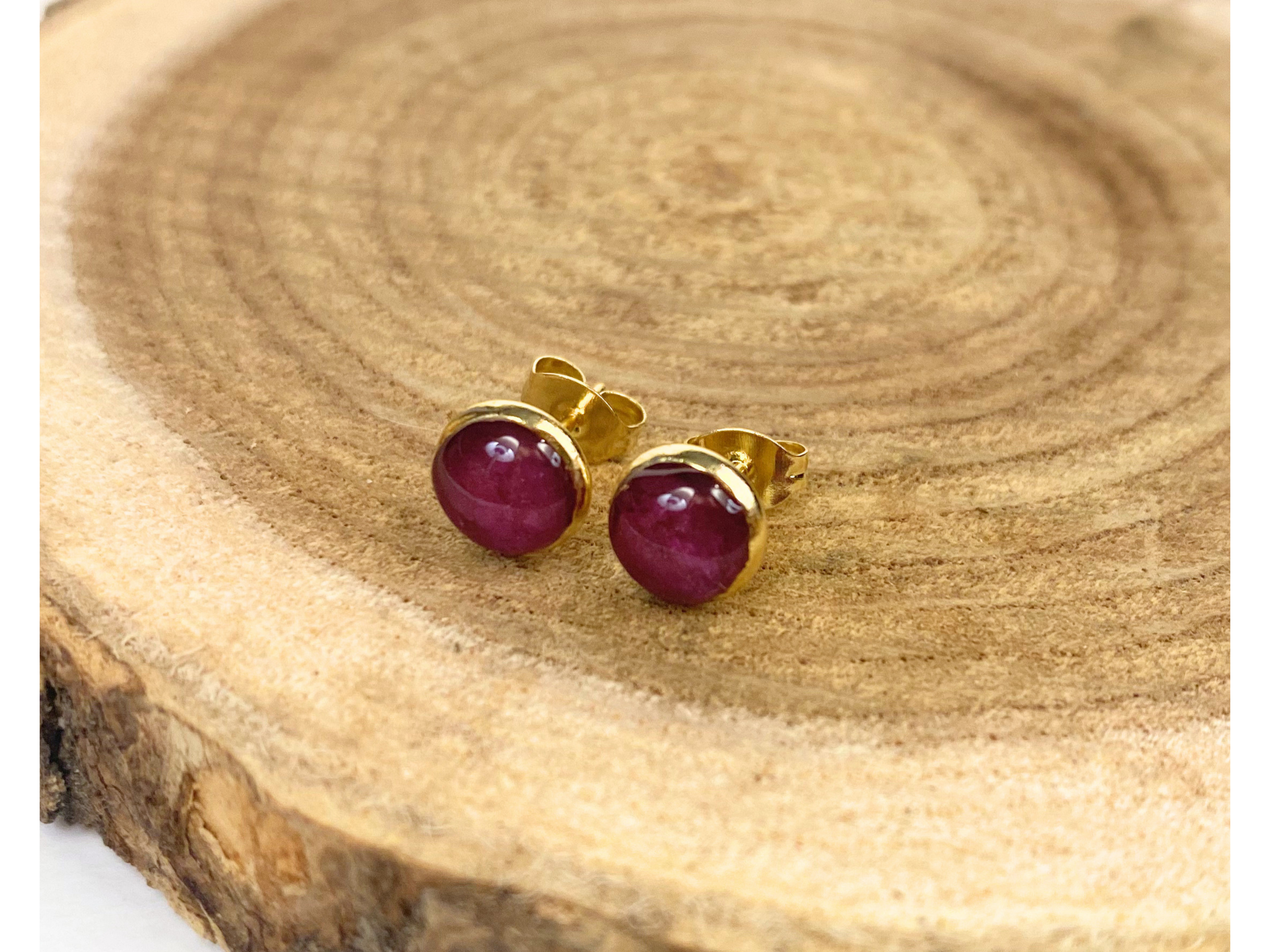 A pair of gold stud earrings containing purple flower petals, displayed on a rustic wood slice background.