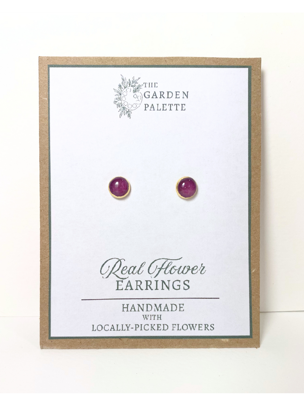 A pair of gold stud earrings containing purple flower petals, mounted on a white display card with a kraft paper border. The card is branded 'The Garden Palette' and labeled 'Real Flower Earrings'.