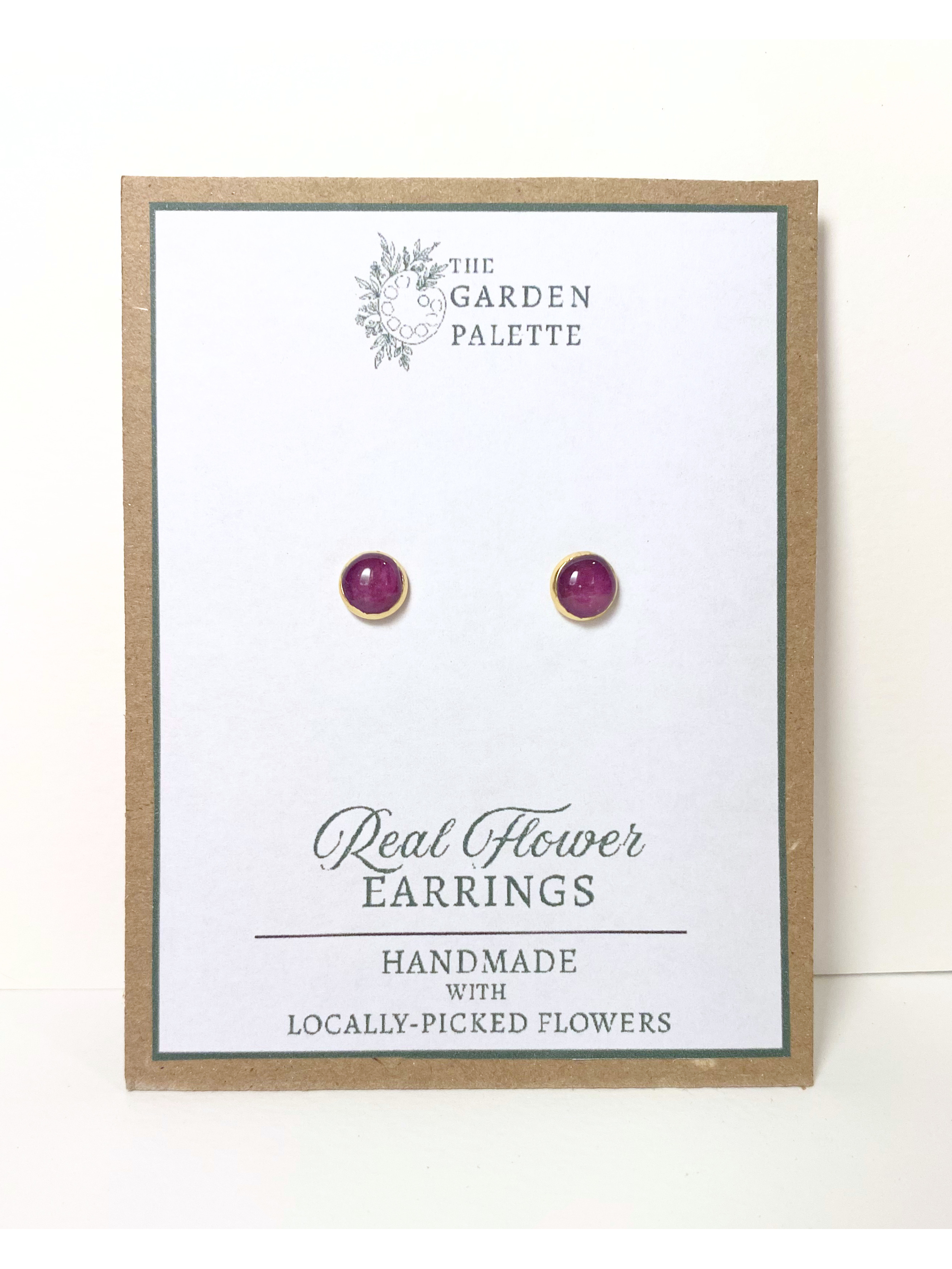A pair of gold stud earrings containing purple flower petals, mounted on a white display card with a kraft paper border. The card is branded 'The Garden Palette' and labeled 'Real Flower Earrings'.