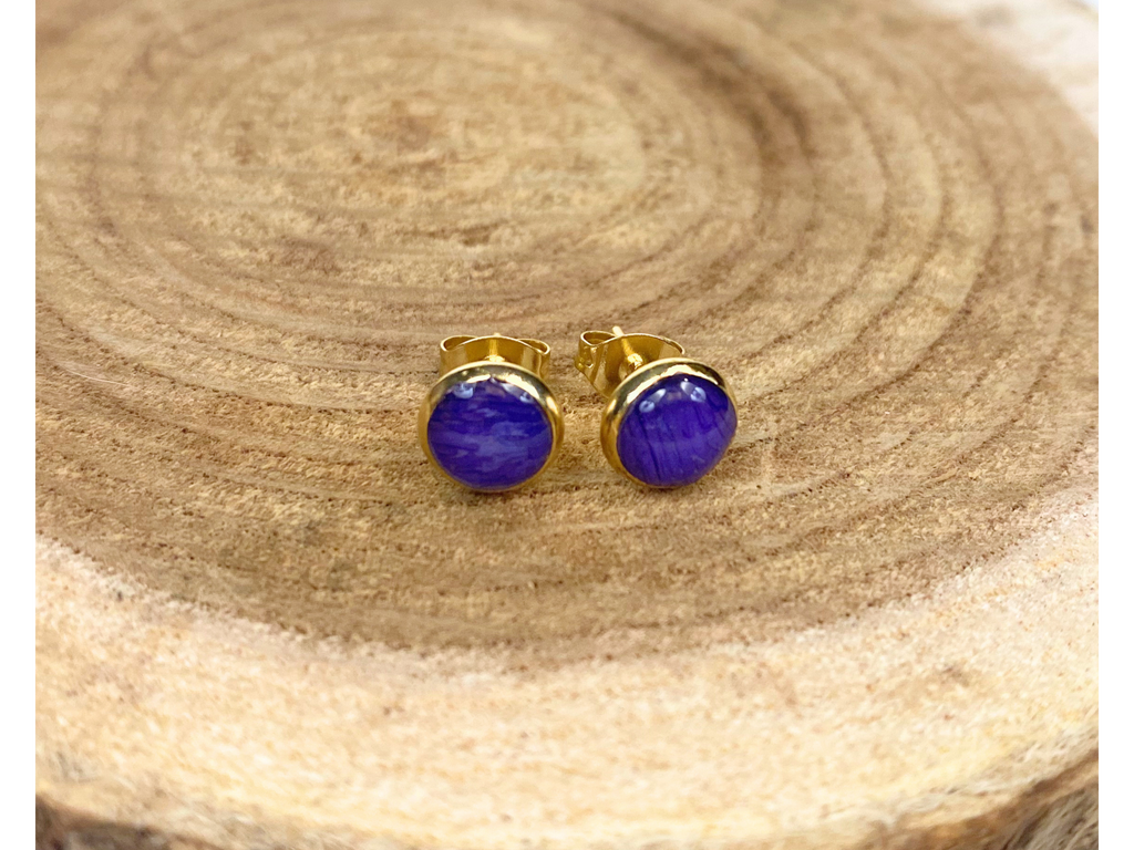 A pair of round gold stud earrings containing blue flower petals, displayed on a textured wood slice background.