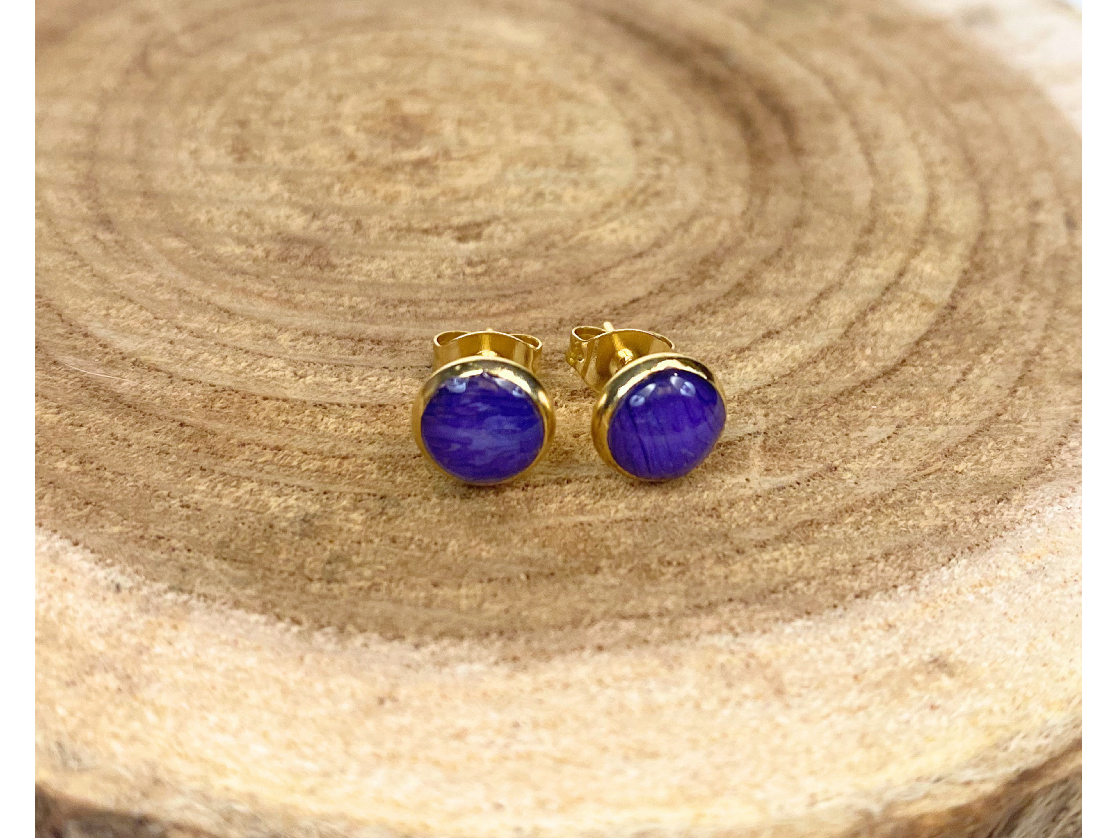 A pair of round gold stud earrings containing blue flower petals, displayed on a textured wood slice background.