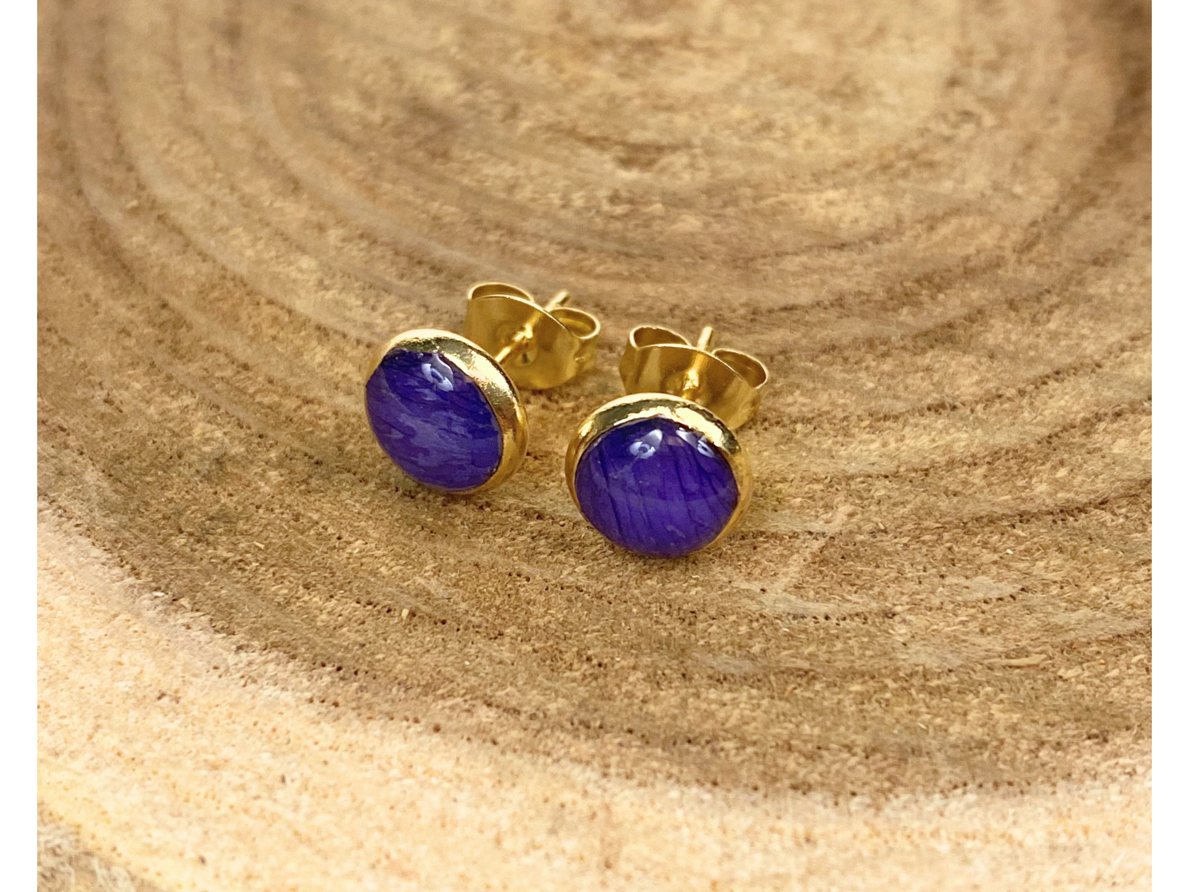 A pair of round gold stud earrings containing blue flower petals, displayed on a textured wood slice background.