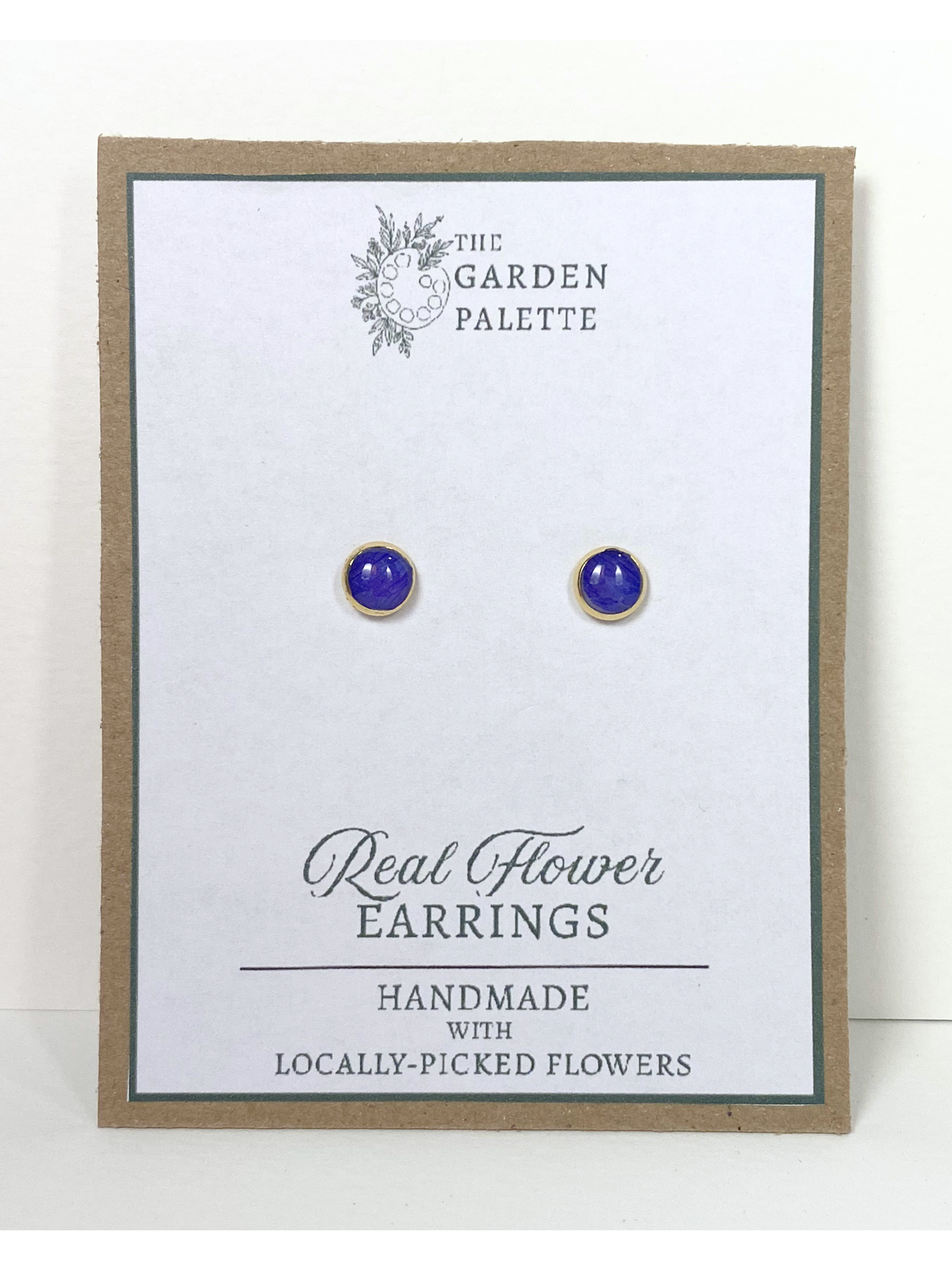 A pair of small stud earrings with deep blue flower petals in gold settings, mounted on a white display card with a kraft paper border. The card is branded 'The Garden Palette' and reads 'Real Flower Earrings'.