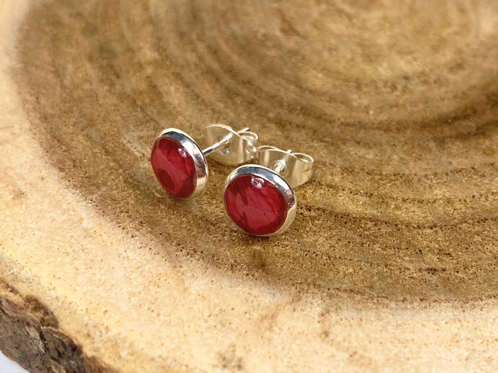 A pair of round silver stud earrings containing deep pink flower petals, displayed on a natural wood slice background.