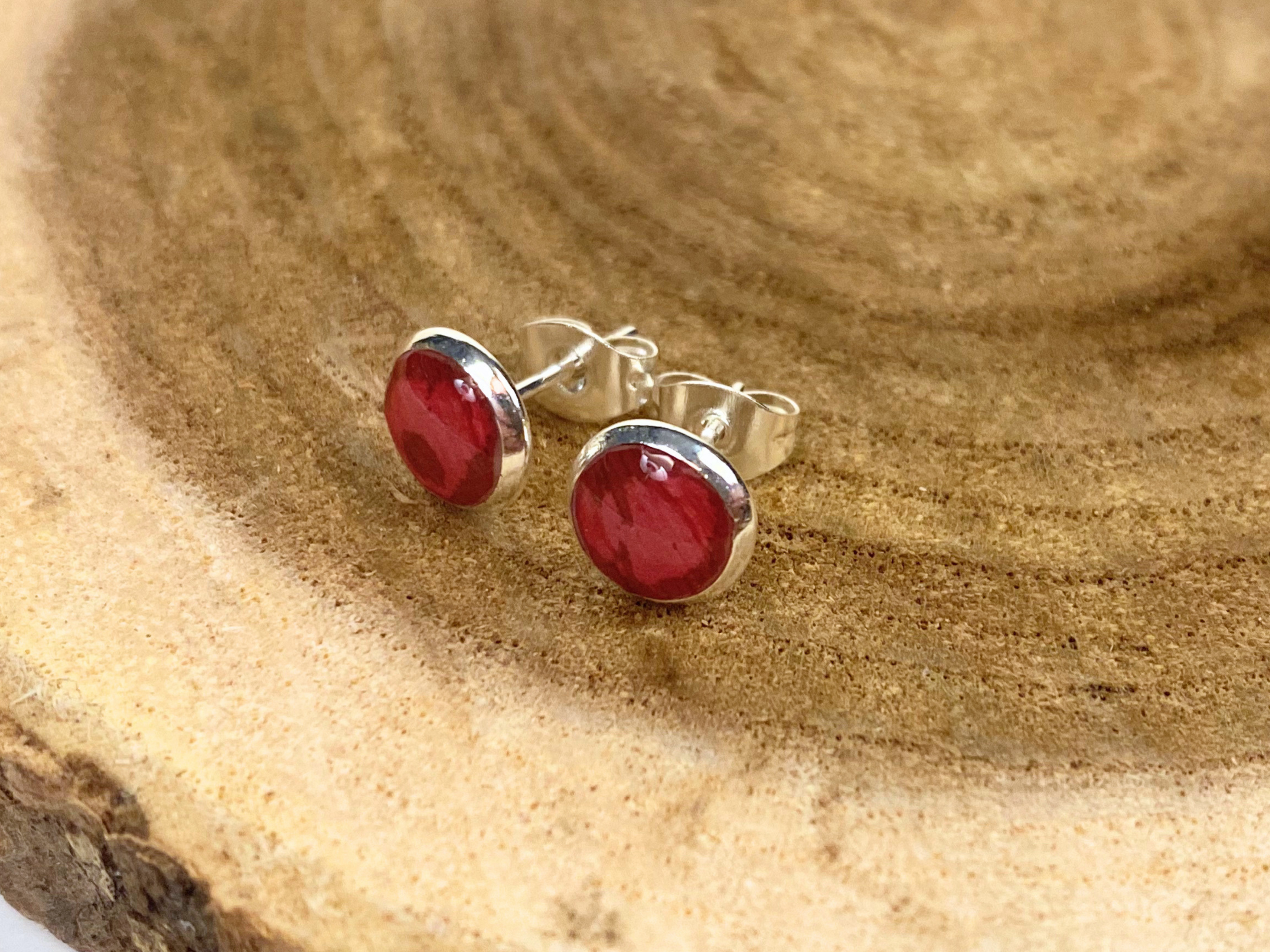 A pair of round silver stud earrings containing deep pink flower petals, displayed on a natural wood slice background.