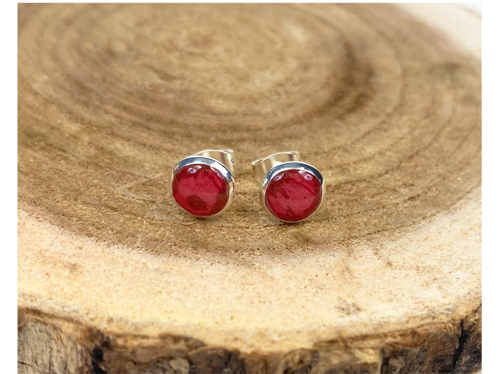 A pair of round silver stud earrings containing deep pink flower petals, displayed on a natural wood slice background.