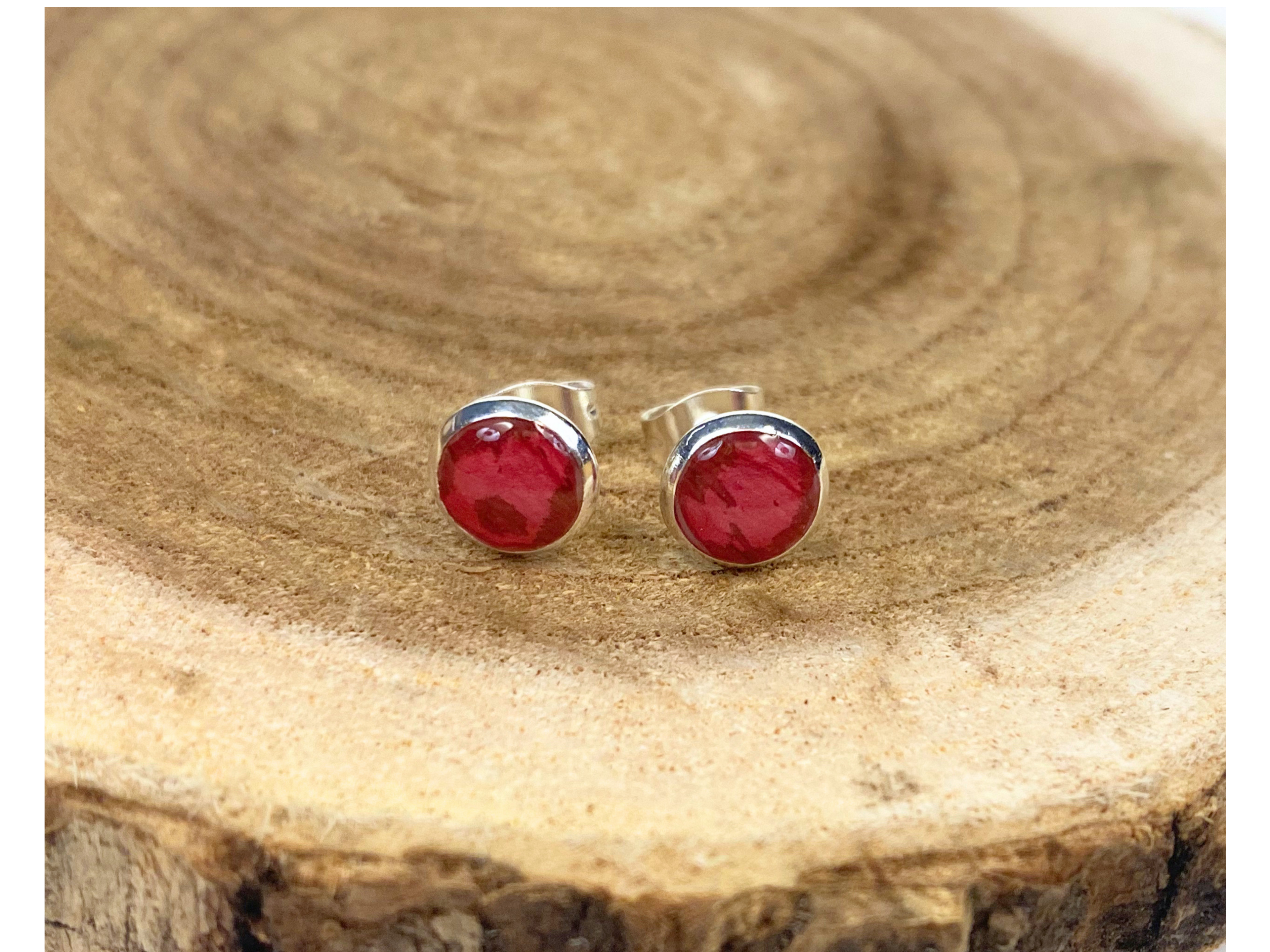 A pair of round silver stud earrings containing deep pink flower petals, displayed on a natural wood slice background.