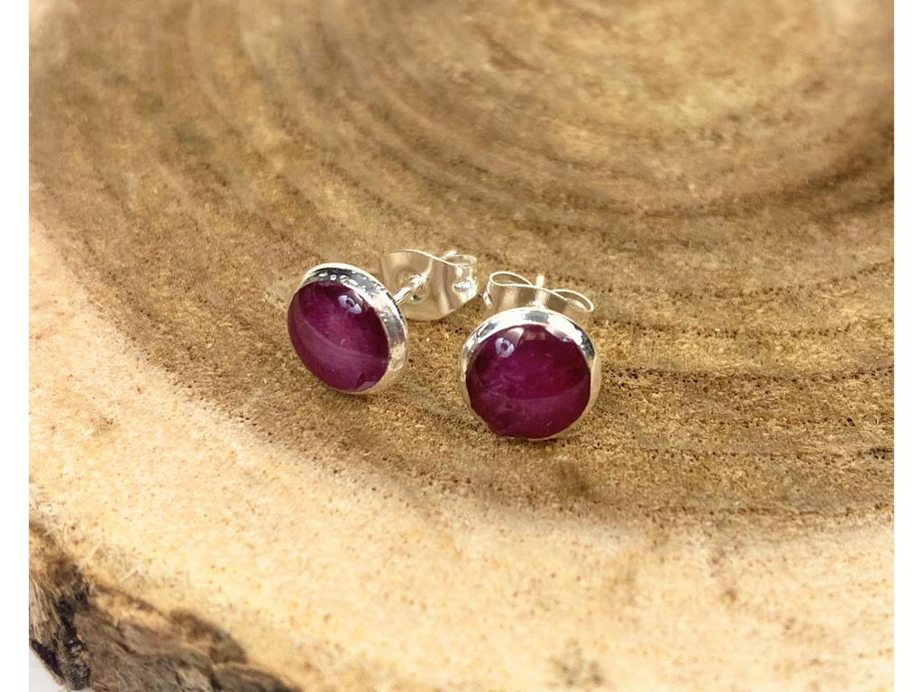 A pair of round silver stud earrings containing purple flower petals, displayed on a rustic wood slice background.