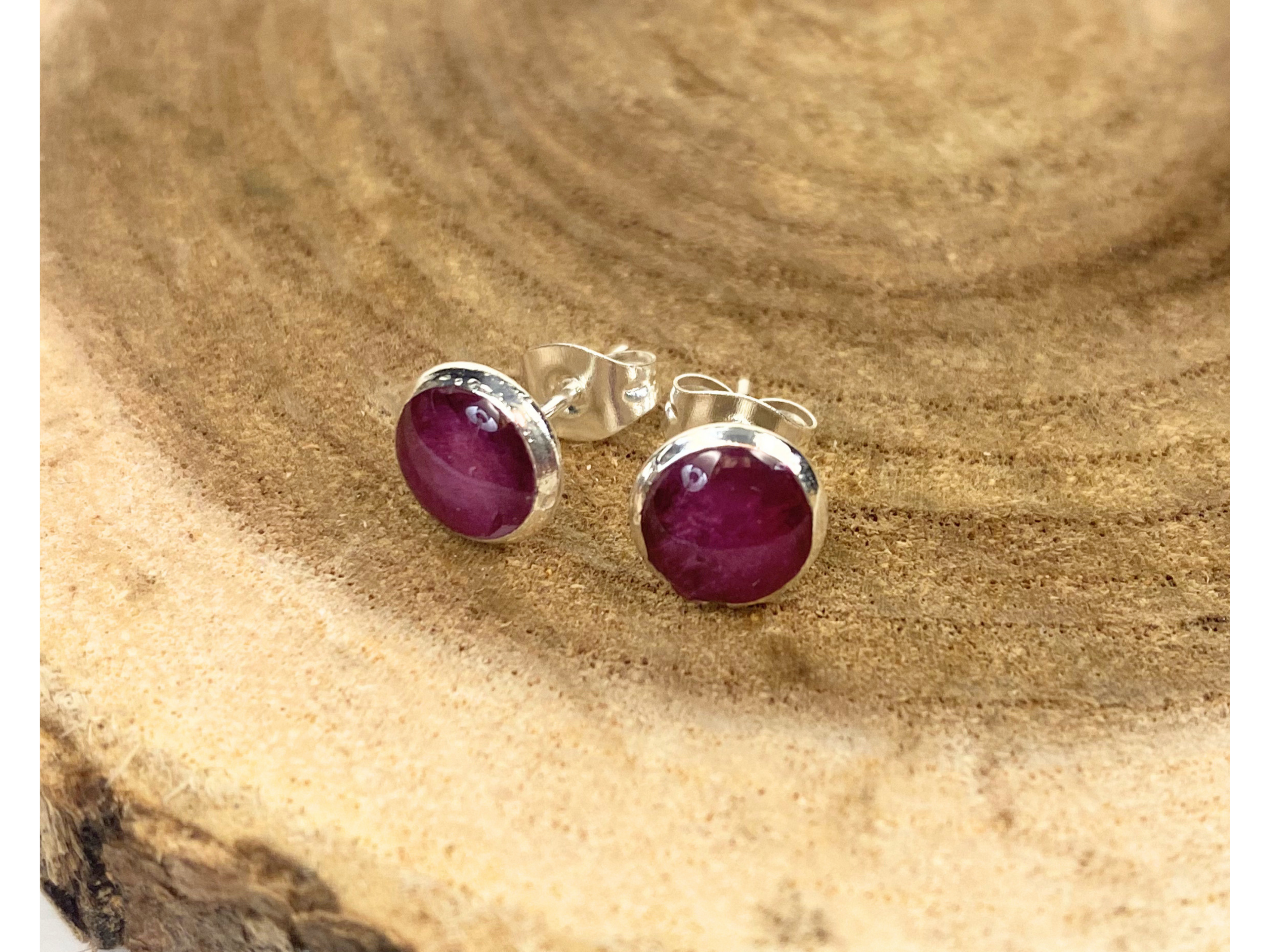 A pair of round silver stud earrings containing purple flower petals, displayed on a rustic wood slice background.