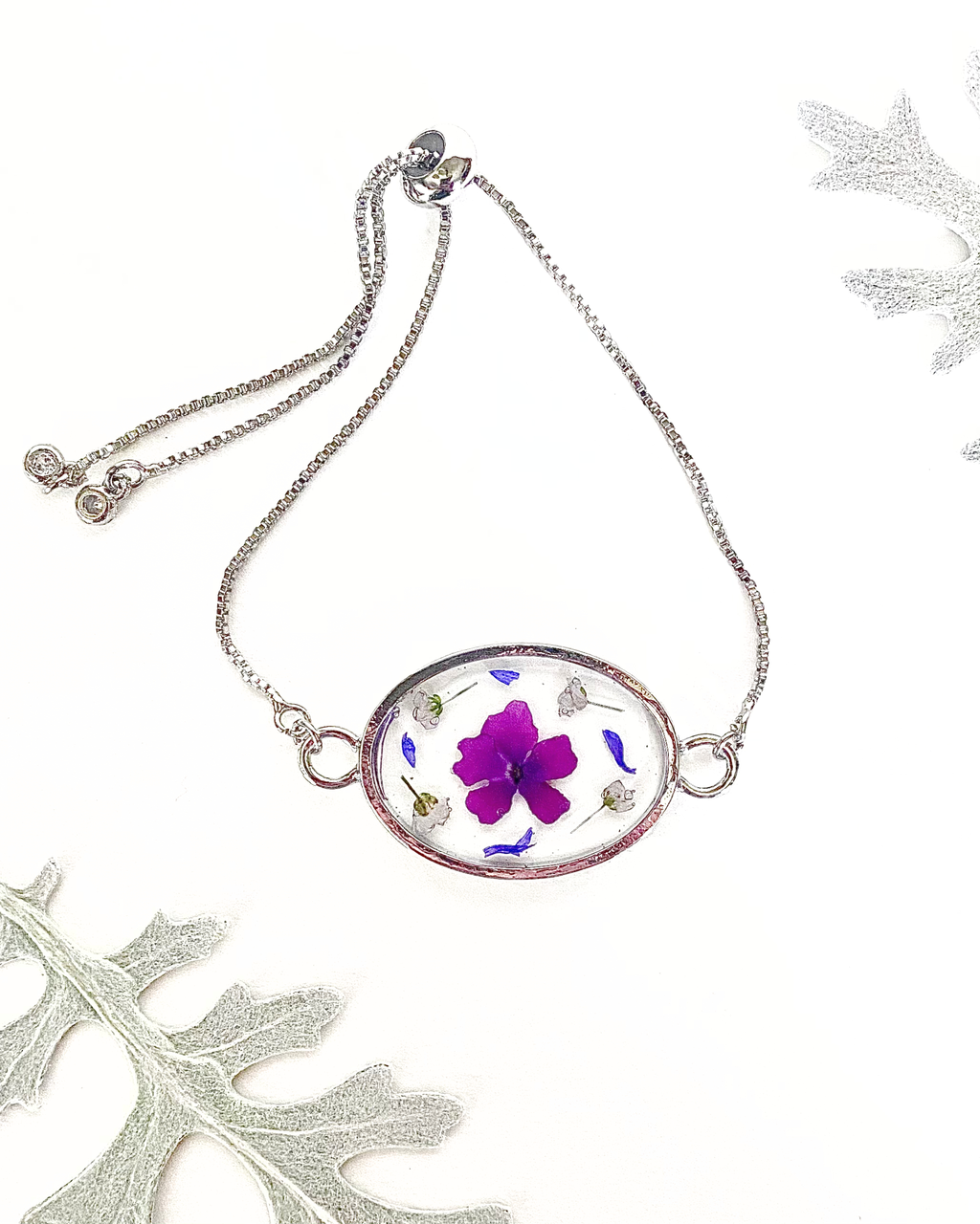 A silver oval pendant bracelet featuring a preserved purple flower and small botanical elements inside clear resin, displayed on a white background with decorative leaves.