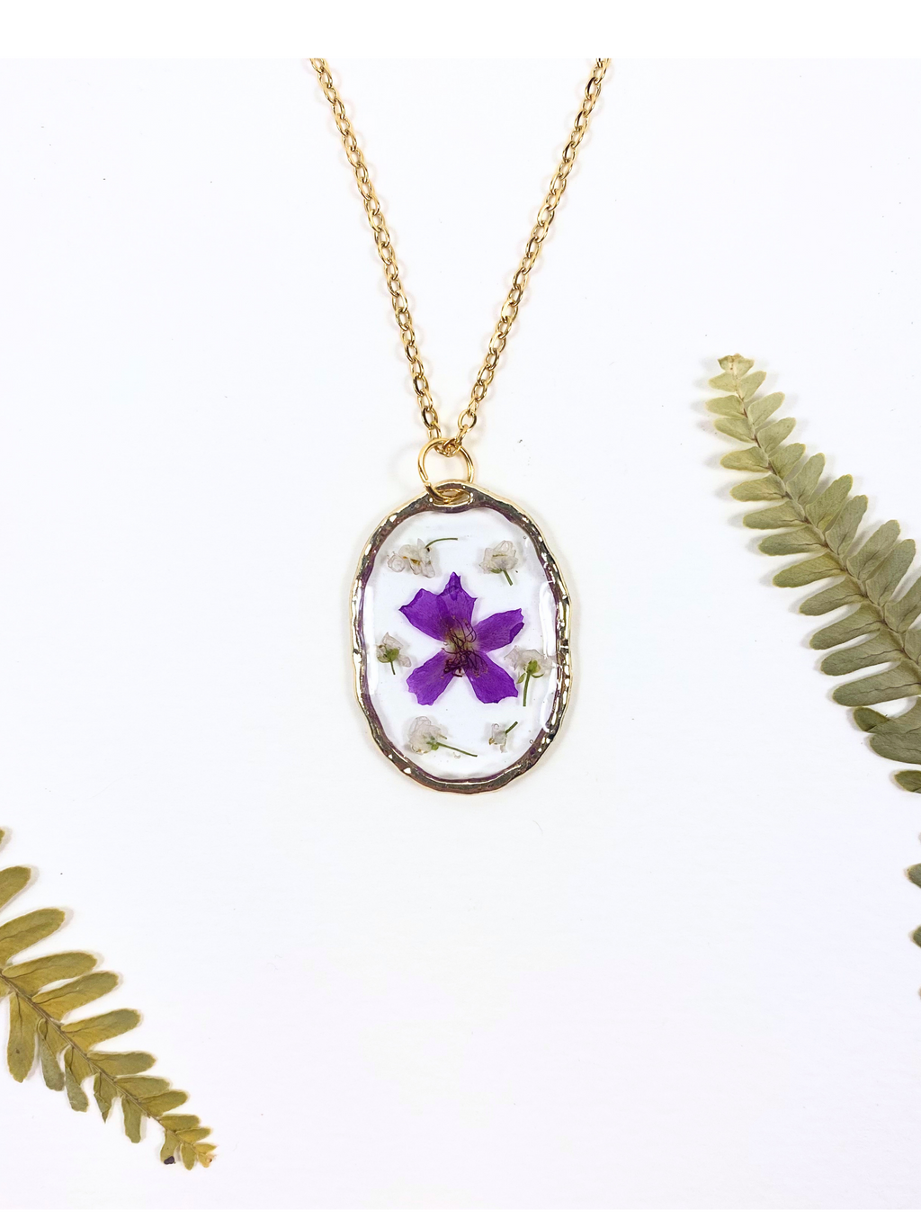 A gold chain necklace with an oval pendant featuring a purple flower and small white blossoms pressed inside a clear resin frame, styled with dried green fern leaves on a white background.