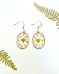 A pair of gold-tone dangle earrings with oval clear resin pendants containing dried yellow and white flowers, displayed on a white background with decorative green fern leaves.