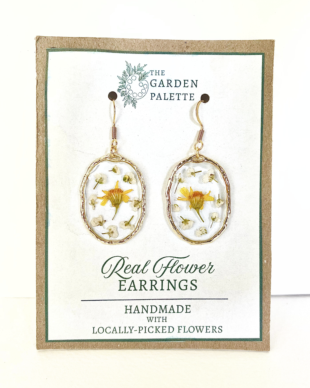 A pair of oval dangling earrings displayed on a branded card. Each earring has a gold-toned frame with a clear dome containing small yellow and white flowers.