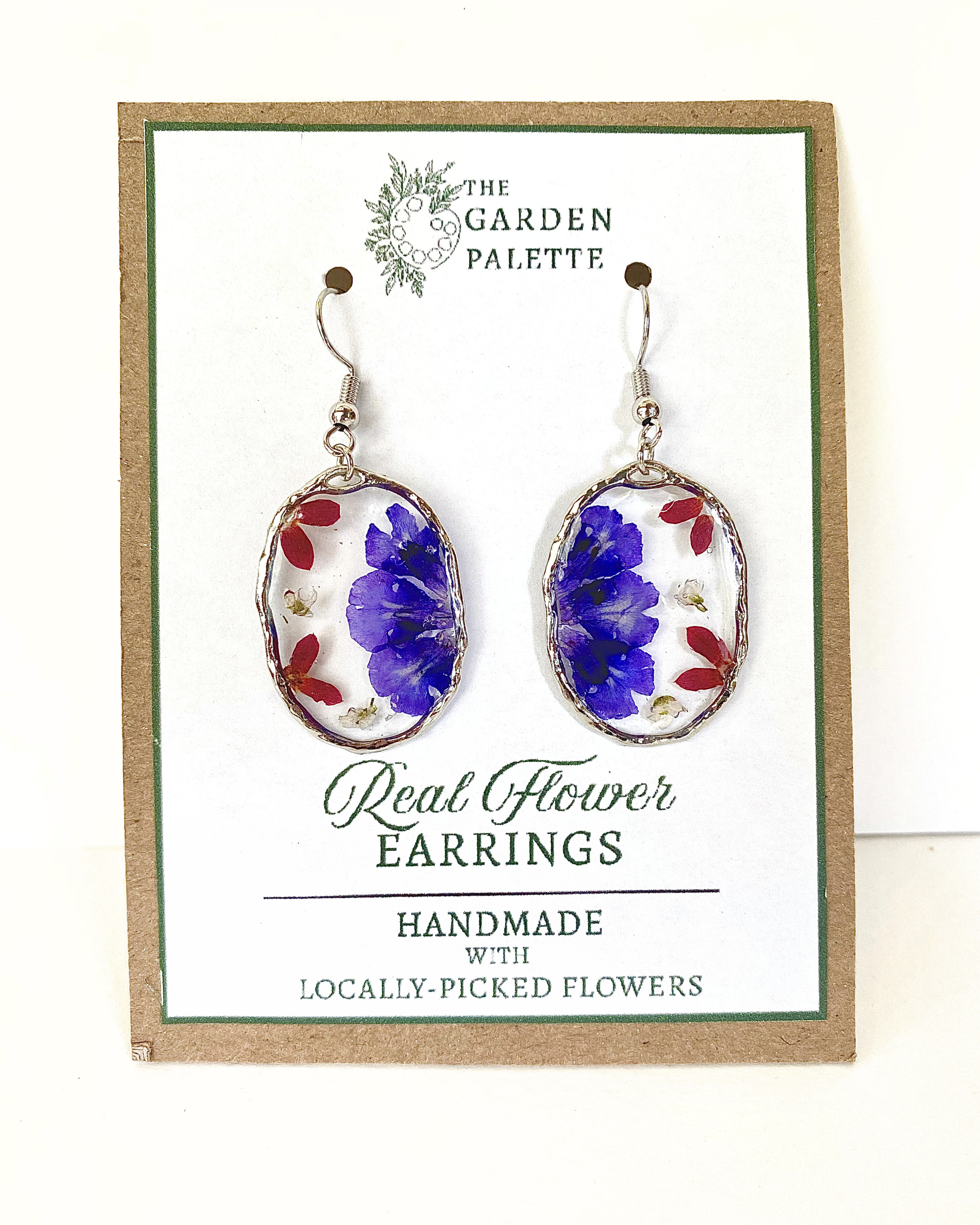 A pair of oval dangling earrings with silver-toned hooks and bezels, featuring pressed blue flowers and small red petals, displayed on a branded card that reads 'The Garden Palette' and 'Real Flower Earrings'.
