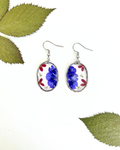 A pair of oval dangling earrings with silver-toned hooks and bezels, featuring pressed blue flowers and small red petals, styled with green leaves on a white surface.