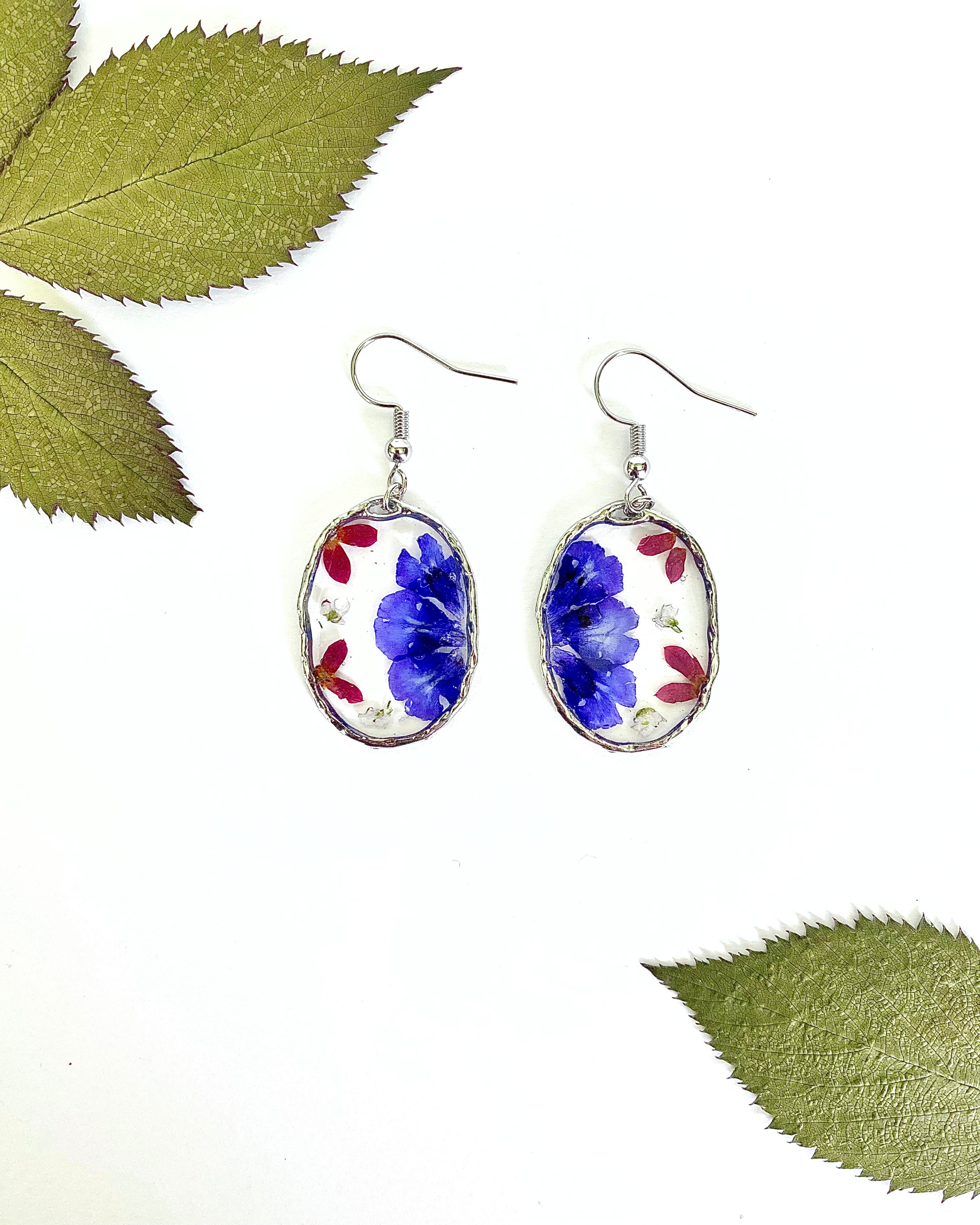 A pair of oval dangling earrings with silver-toned hooks and bezels, featuring pressed blue flowers and small red petals, styled with green leaves on a white surface.