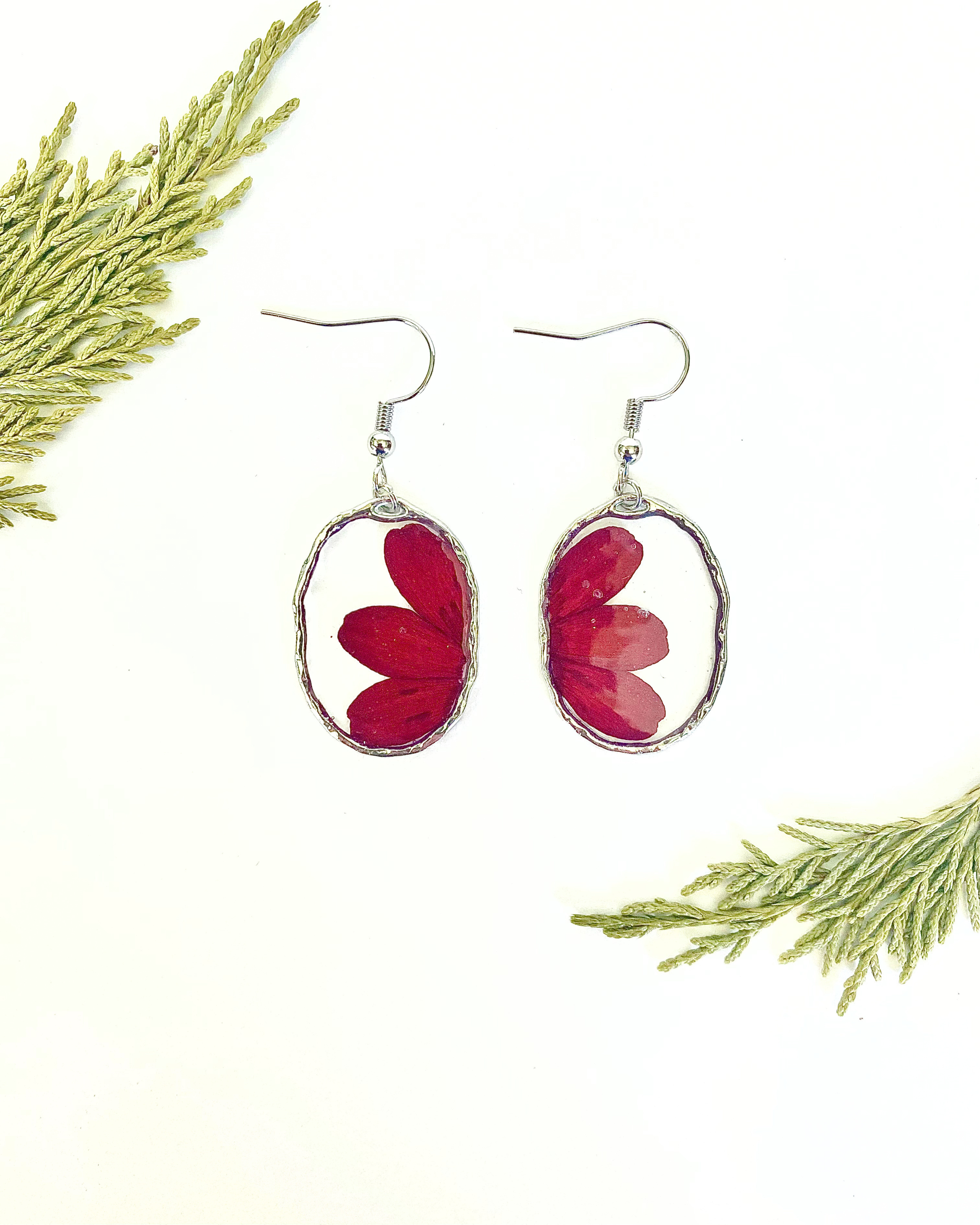 A pair of silver-toned dangle earrings with oval bezel settings containing pressed red flower petals, displayed on a white background with decorative green pine branches in the corners.