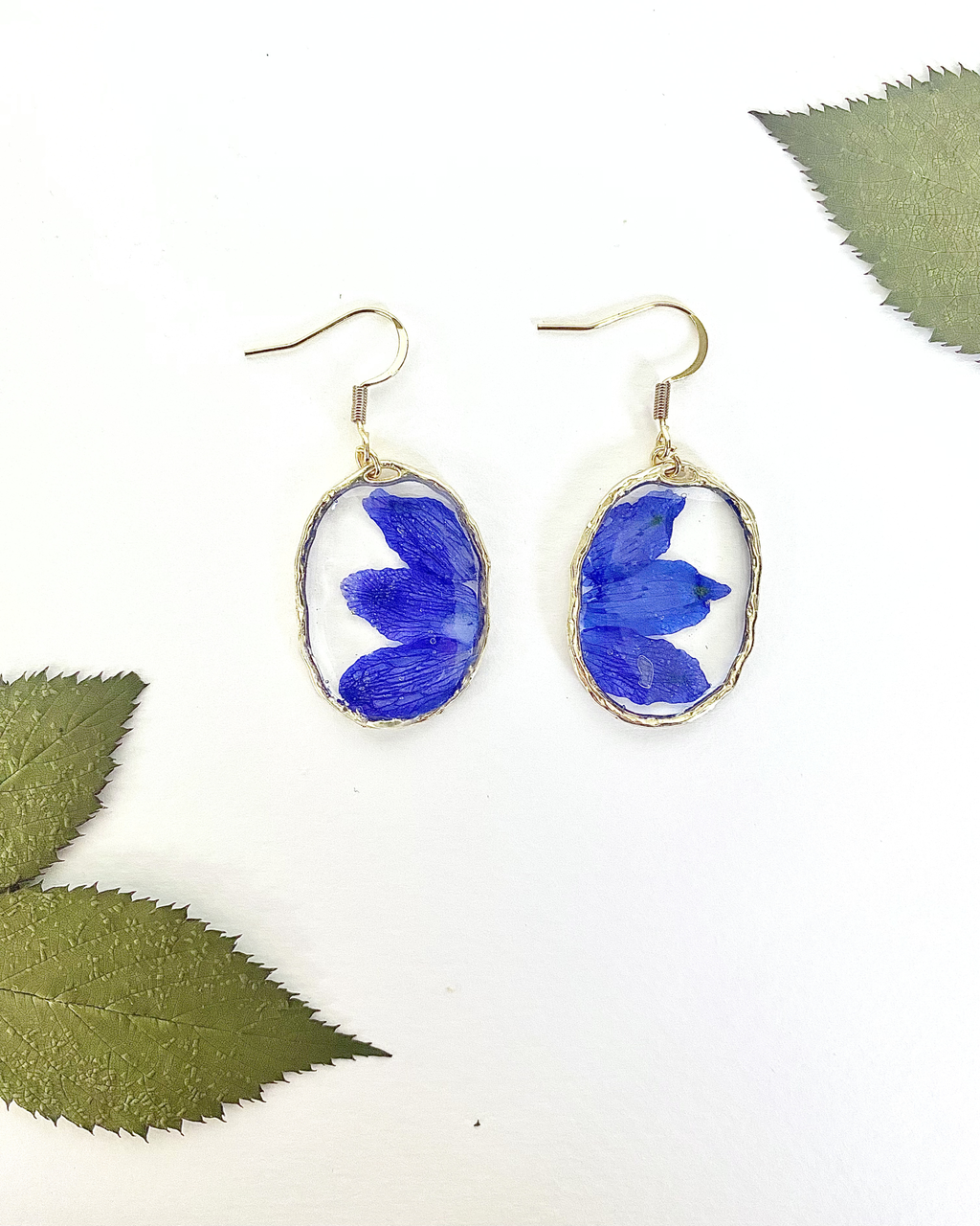 A pair of gold-tone dangle earrings with oval clear resin pendants containing pressed blue flower petals, displayed on a white background with decorative green leaves in the corners.