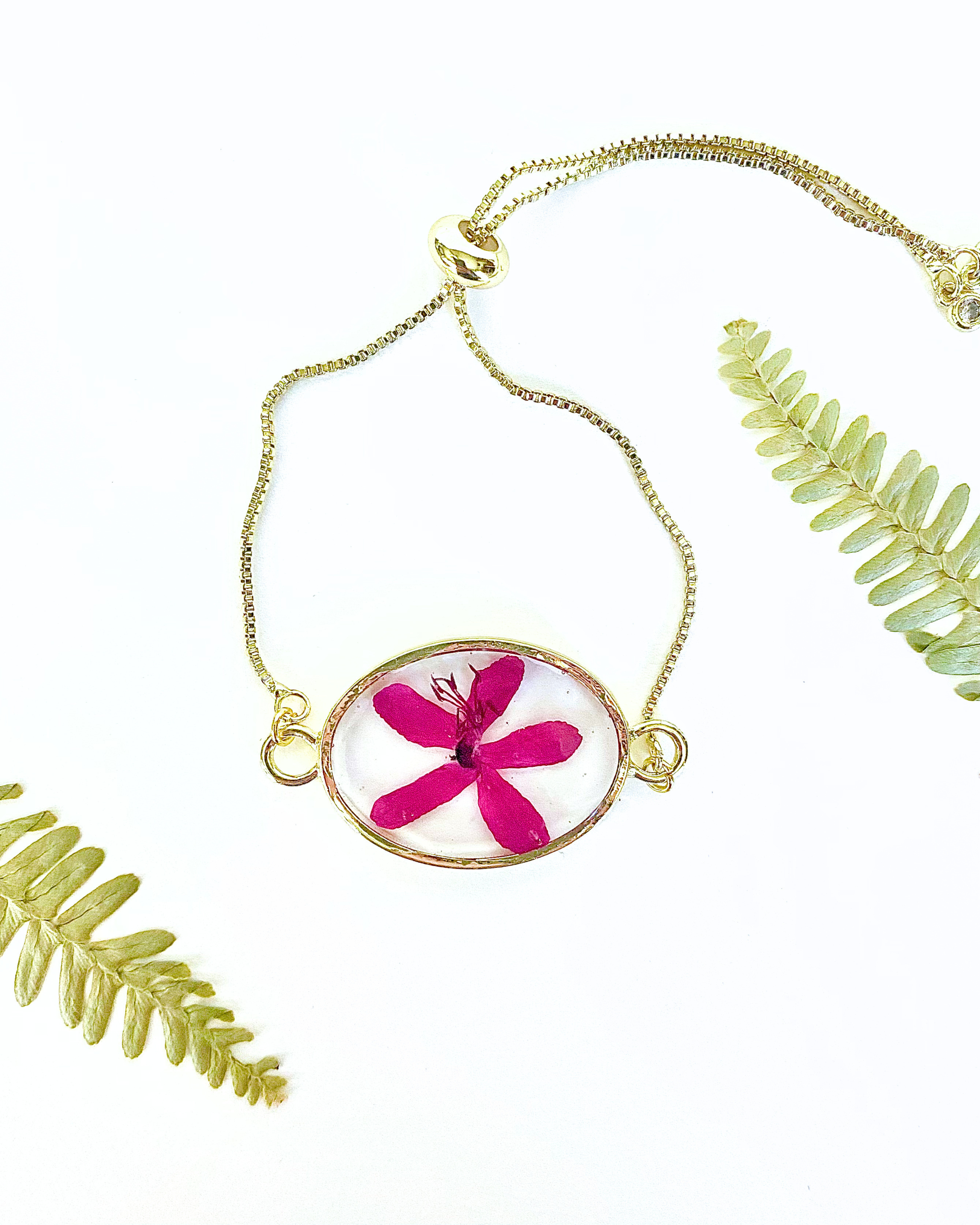 Gold-tone chain bracelet with an oval pendant featuring a bright pink pressed flower in a clear resin, styled with green fern leaves on a white background.