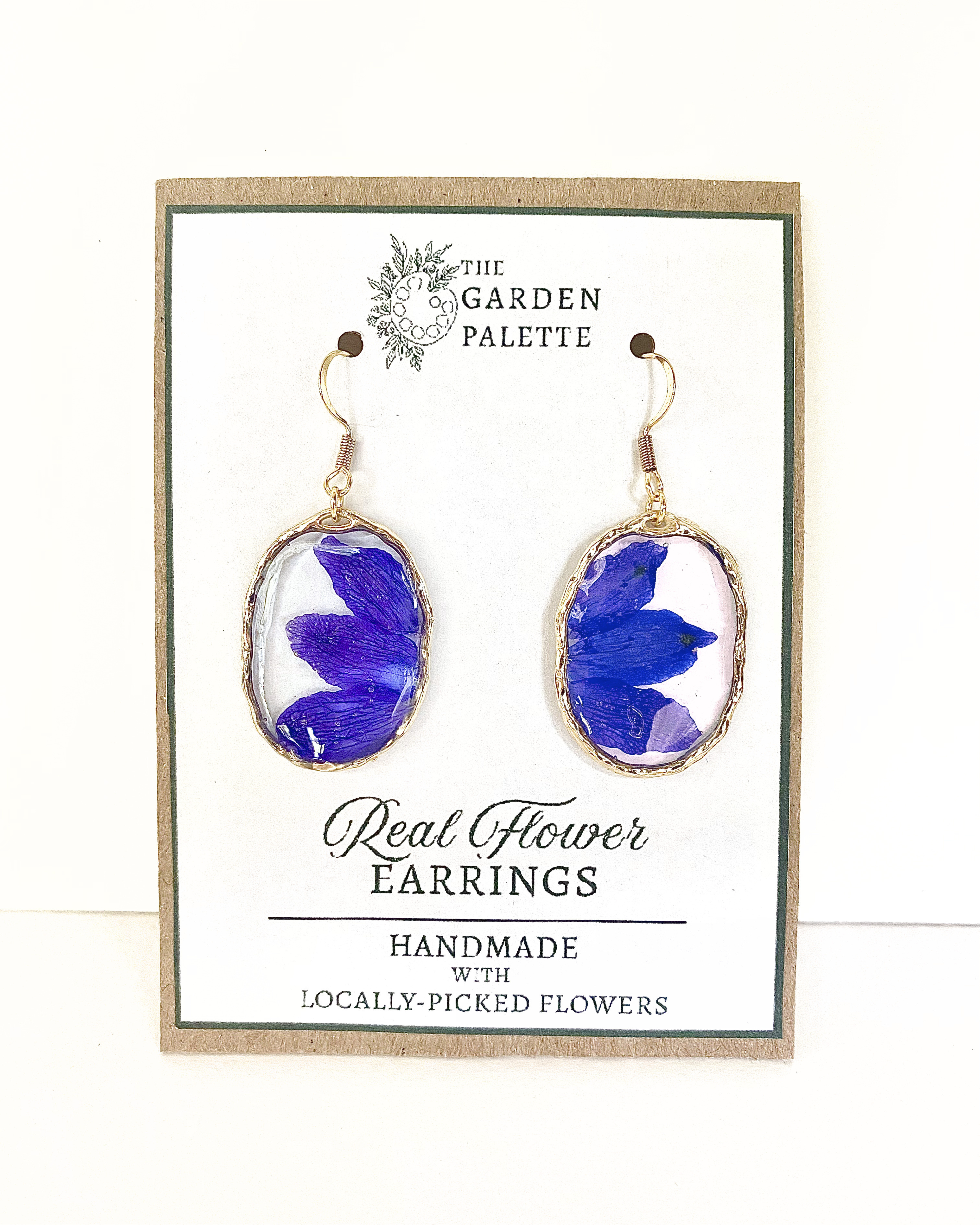 A pair of gold-tone dangle earrings with oval clear resin pendants containing pressed blue flower petals, displayed on a branded card that reads 'The Garden Palette' and 'Real Flower Earrings'.