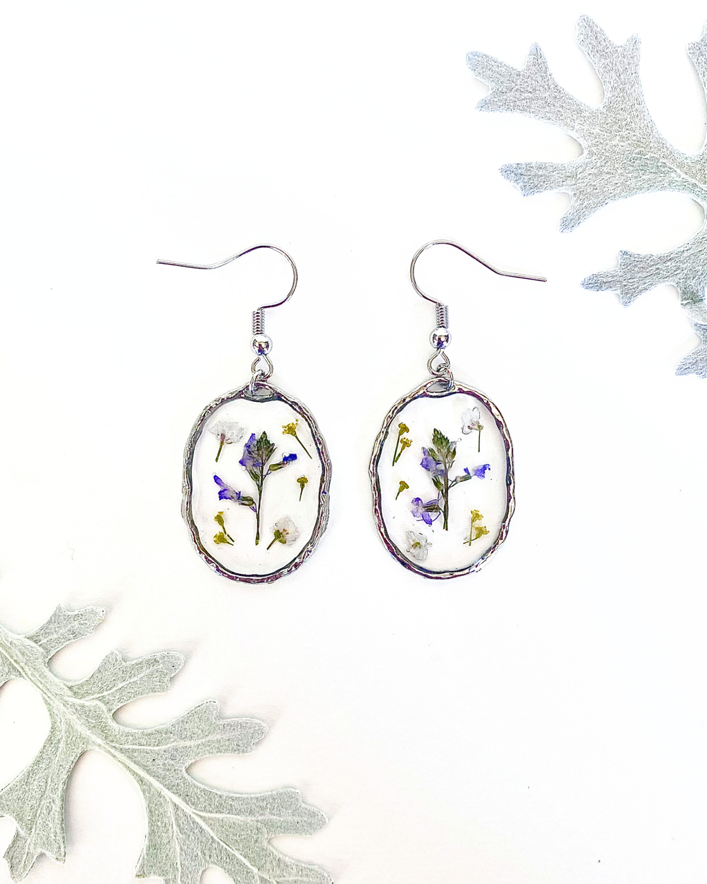 A pair of silver-toned dangle earrings with oval bezel settings containing pressed purple, white, and yellow flowers, displayed on a white background with decorative leaves in the corners.