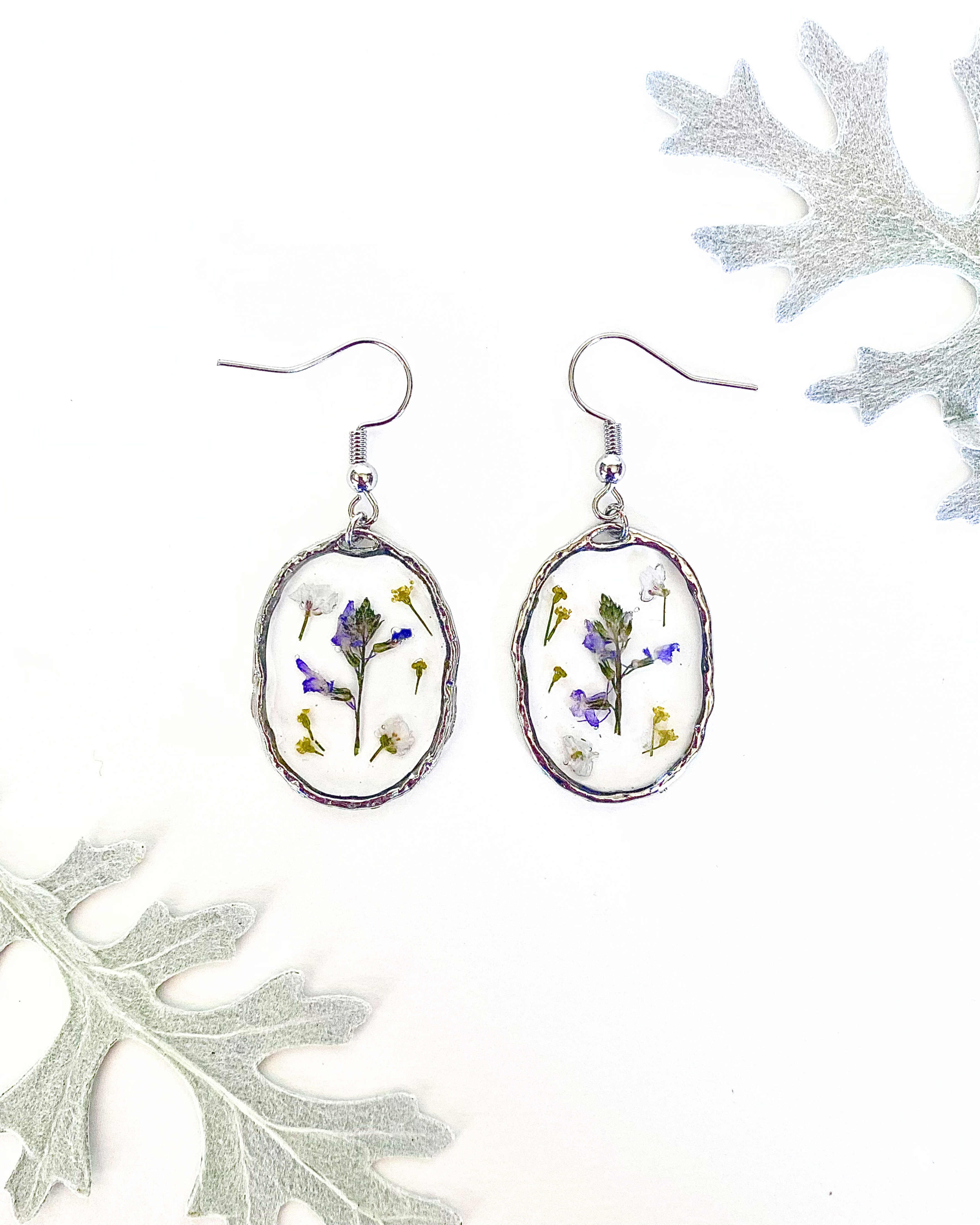 A pair of silver-toned dangle earrings with oval bezel settings containing pressed purple, white, and yellow flowers, displayed on a white background with decorative leaves in the corners.