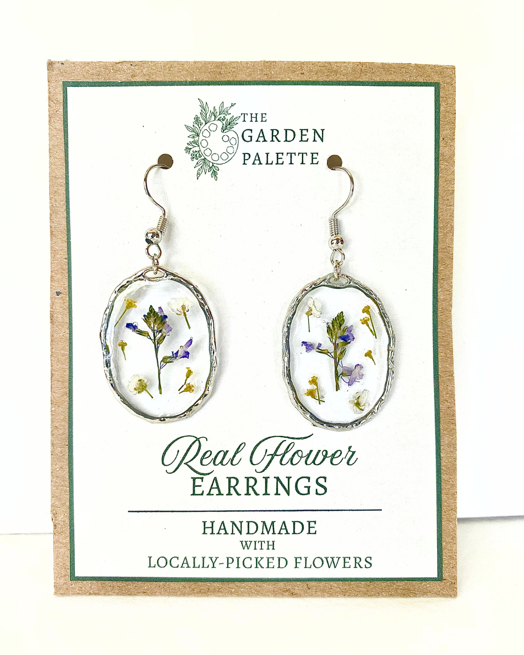 A pair of silver-toned dangle earrings with oval bezel settings containing pressed purple, white, and yellow flowers, displayed on a branded card that reads 'The Garden Palette' and 'Real Flower Earrings'.