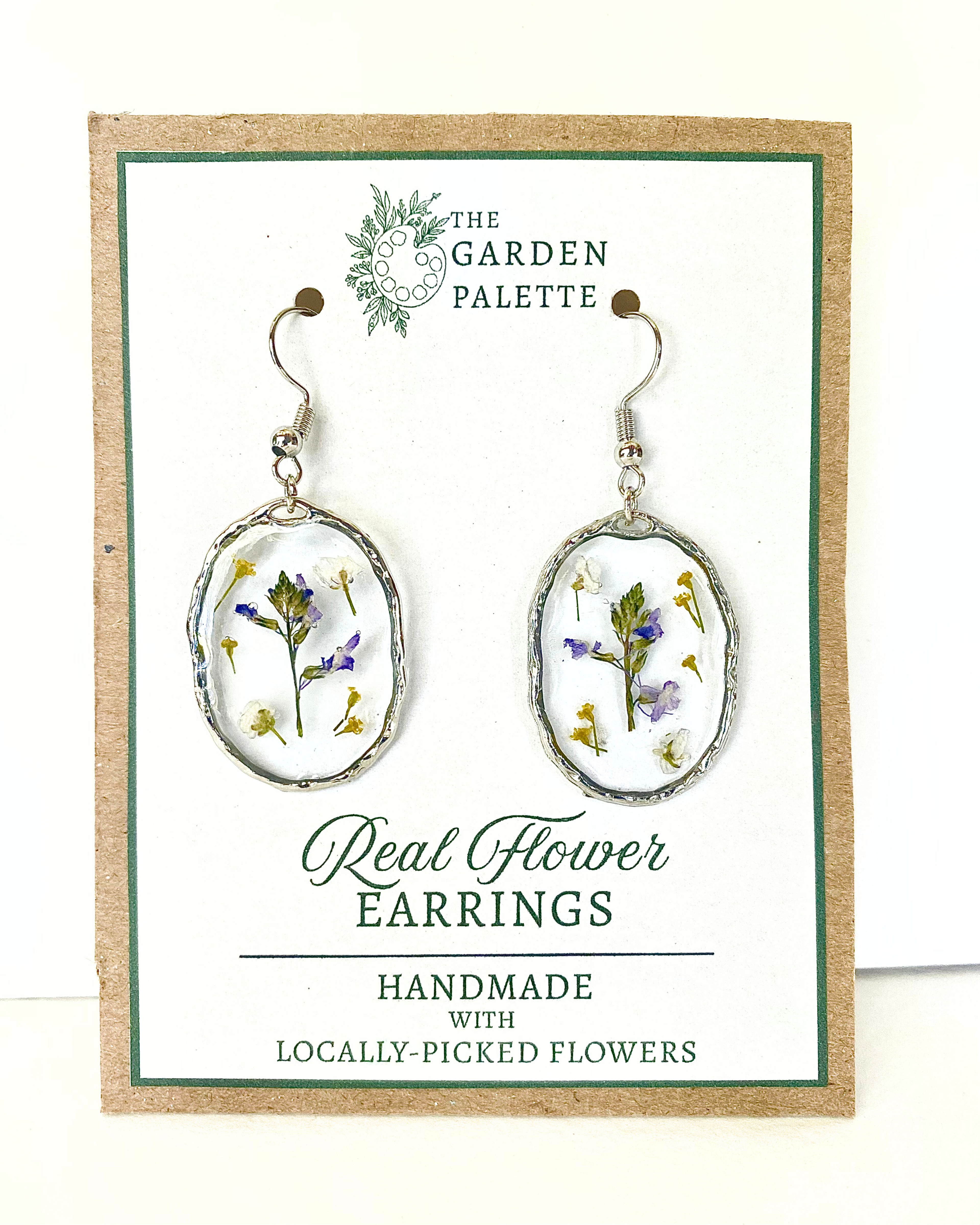 A pair of silver-toned dangle earrings with oval bezel settings containing pressed purple, white, and yellow flowers, displayed on a branded card that reads 'The Garden Palette' and 'Real Flower Earrings'.