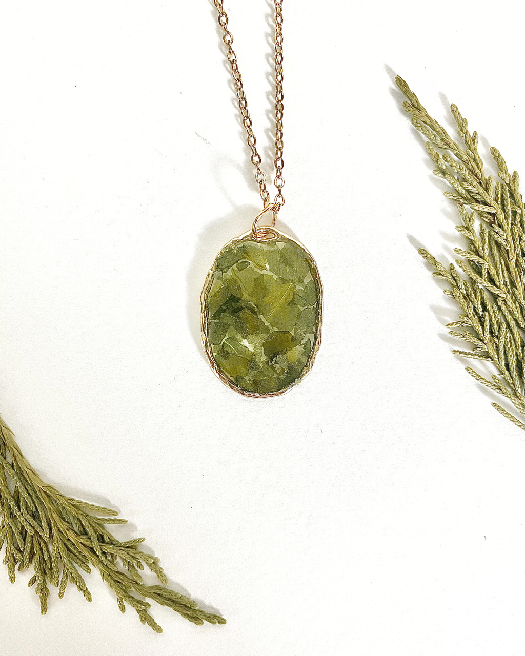 A gold-toned necklace chain with an oval bezel setting containing pressed green leave pieces, displayed on a white background with decorative green pine branches in the corners.