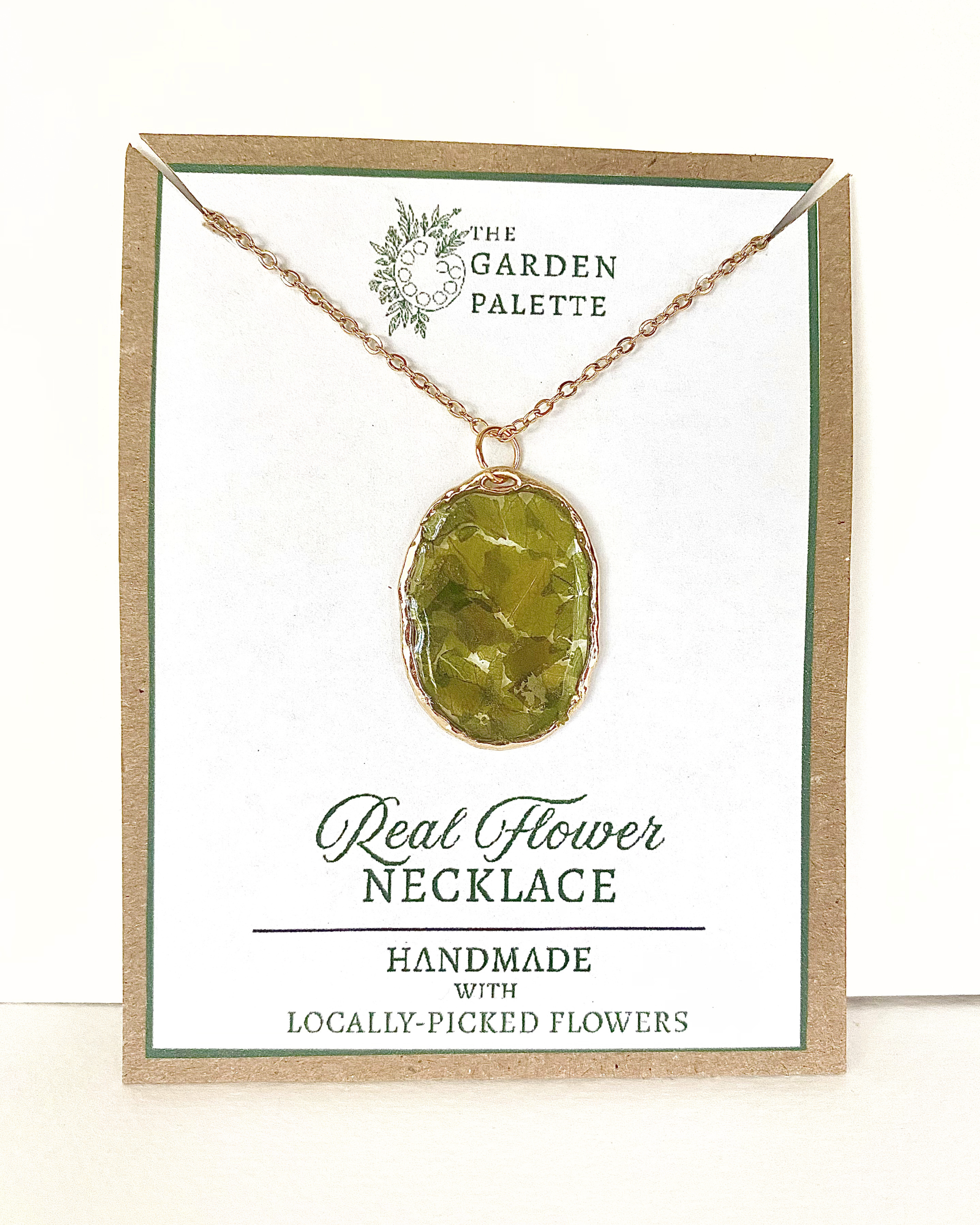 A gold-toned necklace chain with an oval bezel setting containing pressed green leave pieces, displayed on a branded card that reads 'The Garden Palette' and 'Real Flower Necklace'.