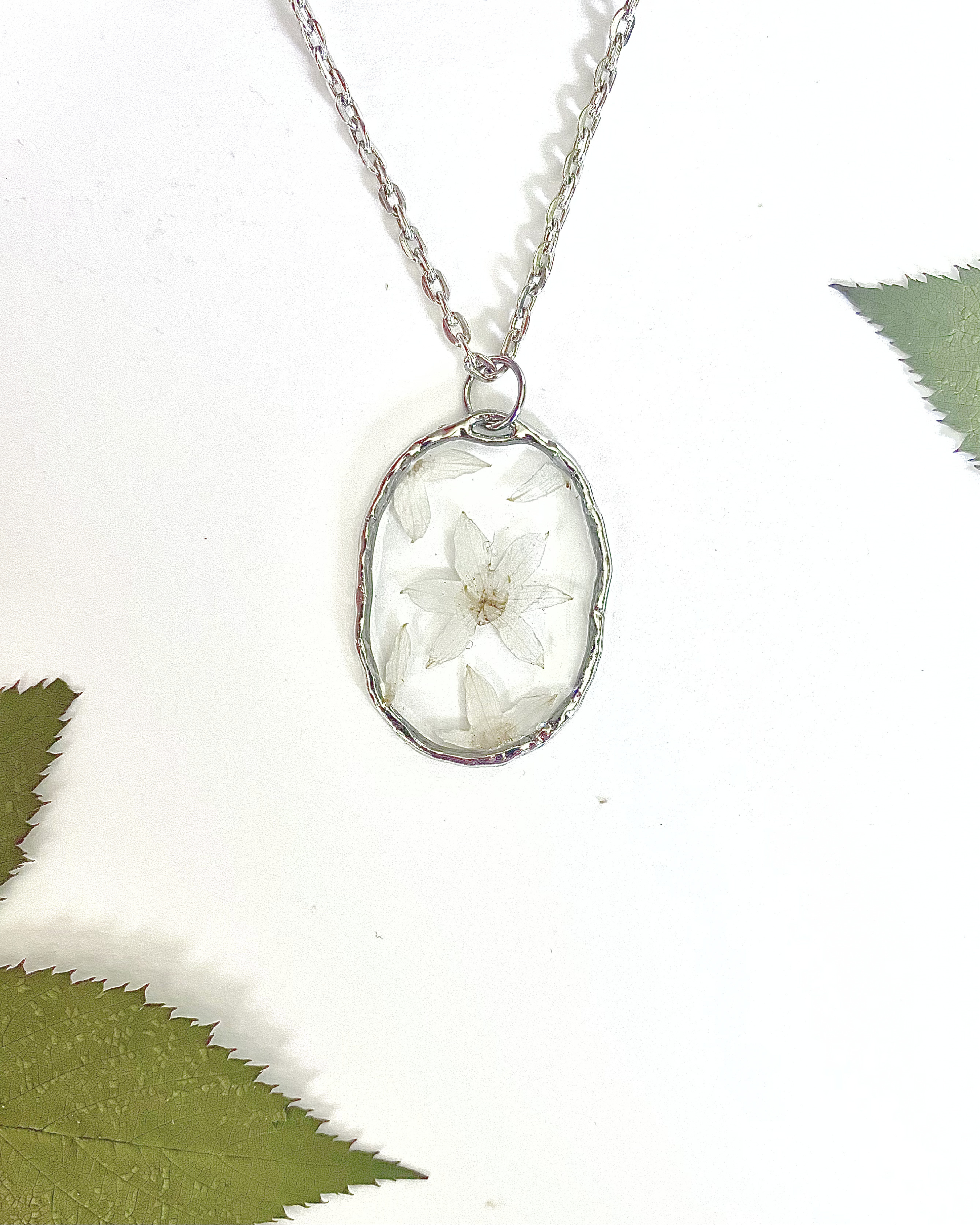 A silver-toned necklace chain with an oval bezel setting containing pressed white flowers, displayed on a white background with decorative green leaves in the corners.