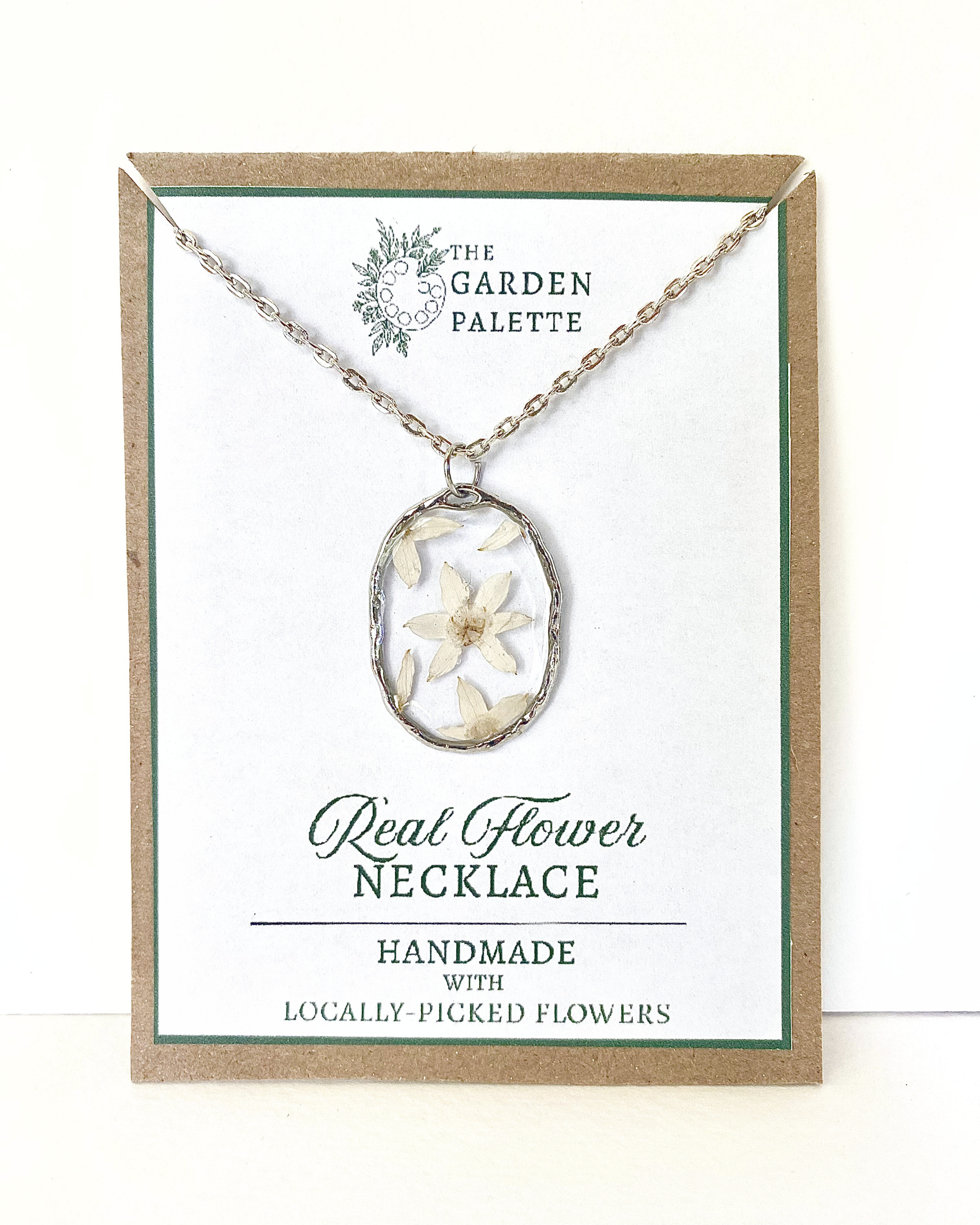 A silver-toned necklace chain with an oval bezel setting containing pressed white flowers, displayed on a branded card that reads 'The Garden Palette' and 'Real Flower Necklace'.