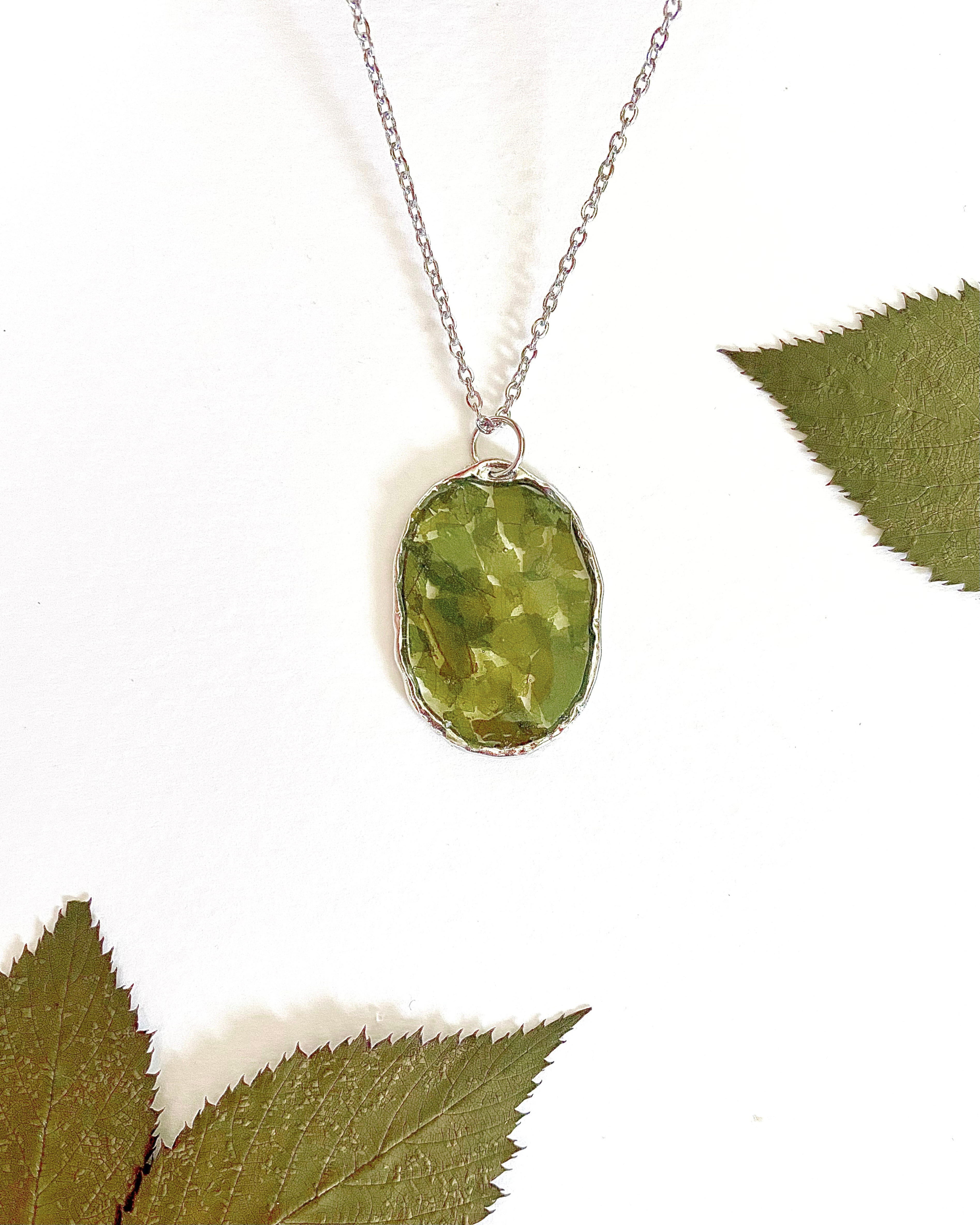 A silver-toned necklace chain with an oval bezel setting containing pressed green leave pieces, displayed on a white background with decorative green leaves in the corners.