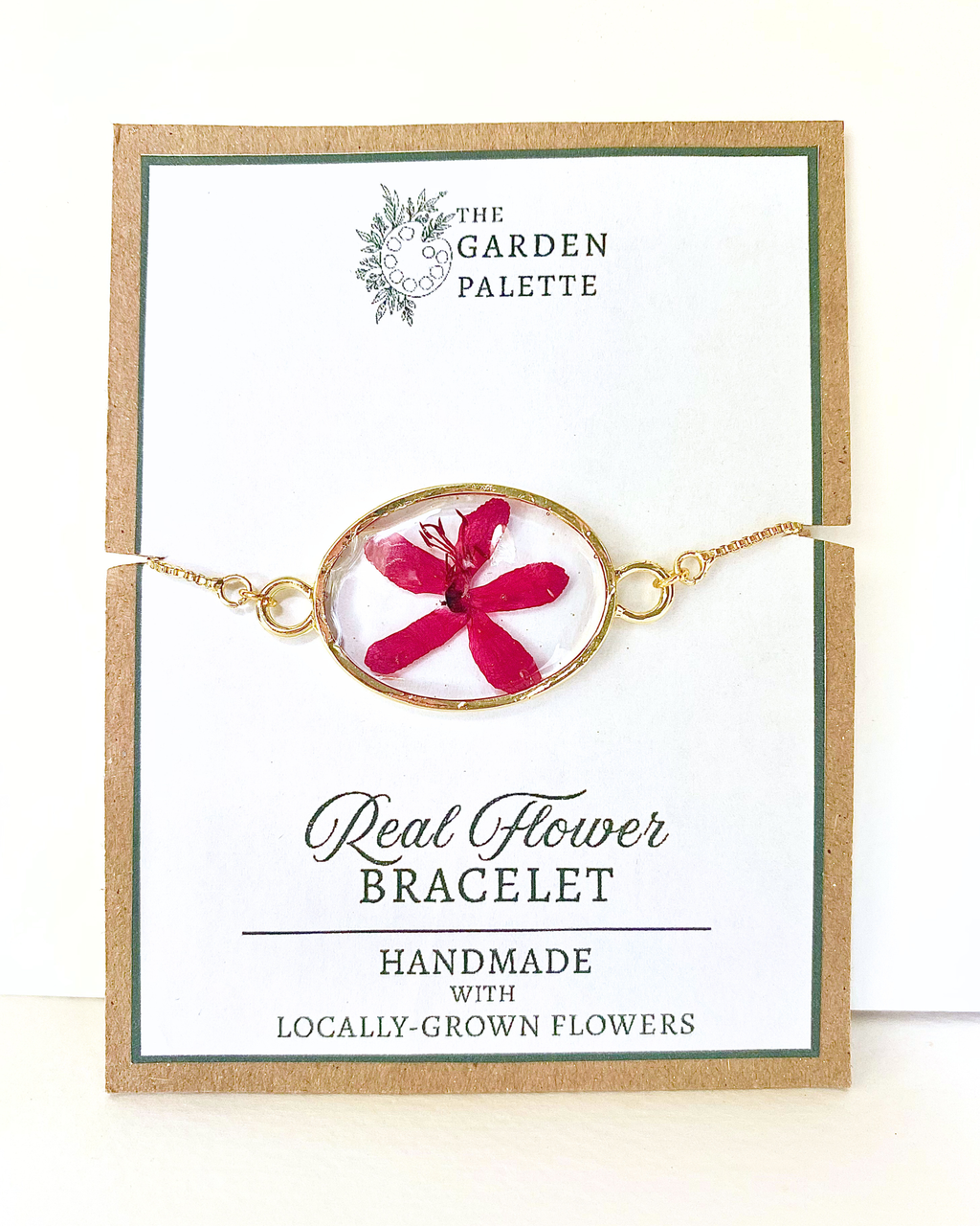 Gold-tone chain bracelet with an oval pendant featuring a bright pink pressed flower in a clear resin, displayed on a branded card that reads 'The Garden Palette' and 'Real Flower Bracelet'.