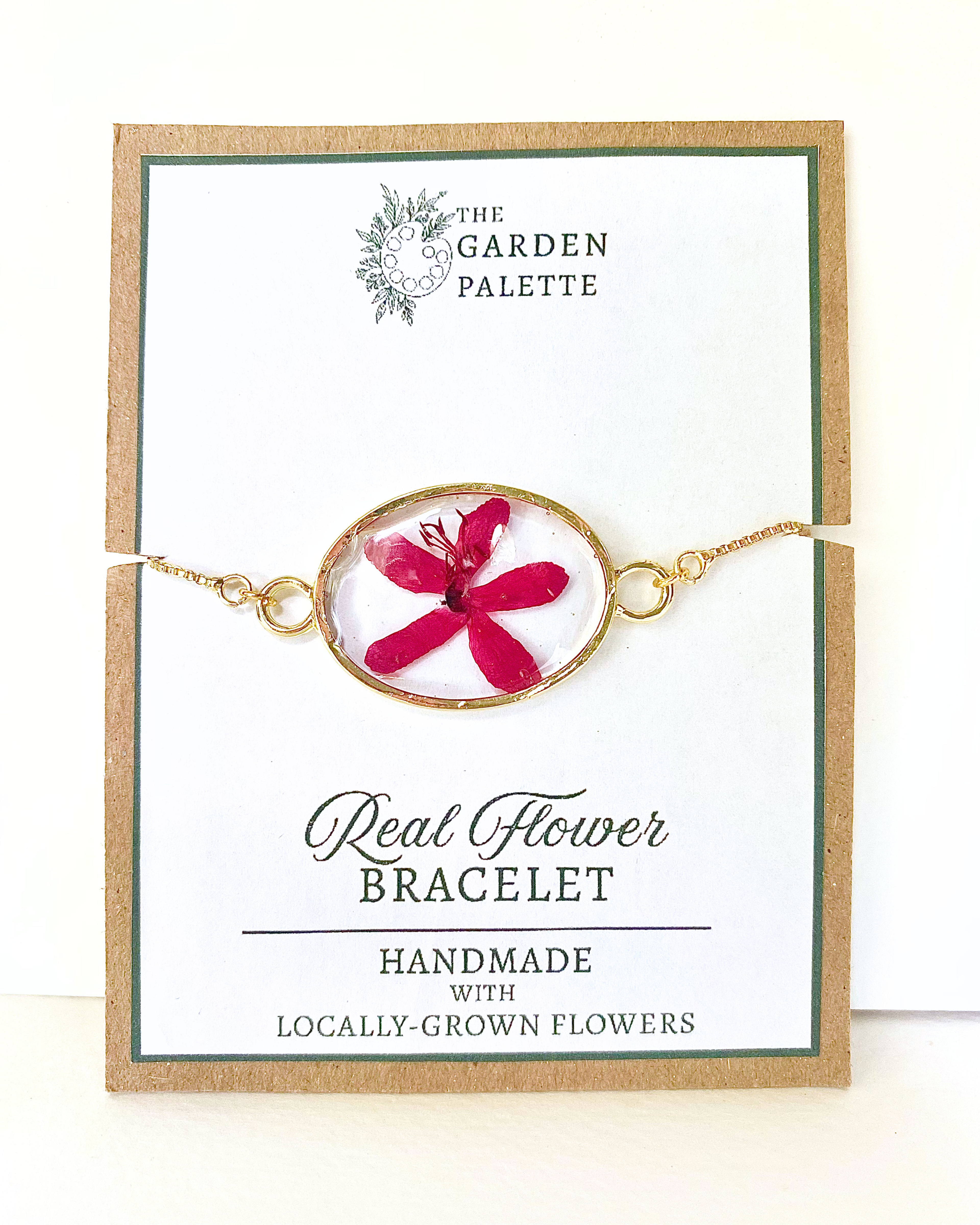 Gold-tone chain bracelet with an oval pendant featuring a bright pink pressed flower in a clear resin, displayed on a branded card that reads 'The Garden Palette' and 'Real Flower Bracelet'.