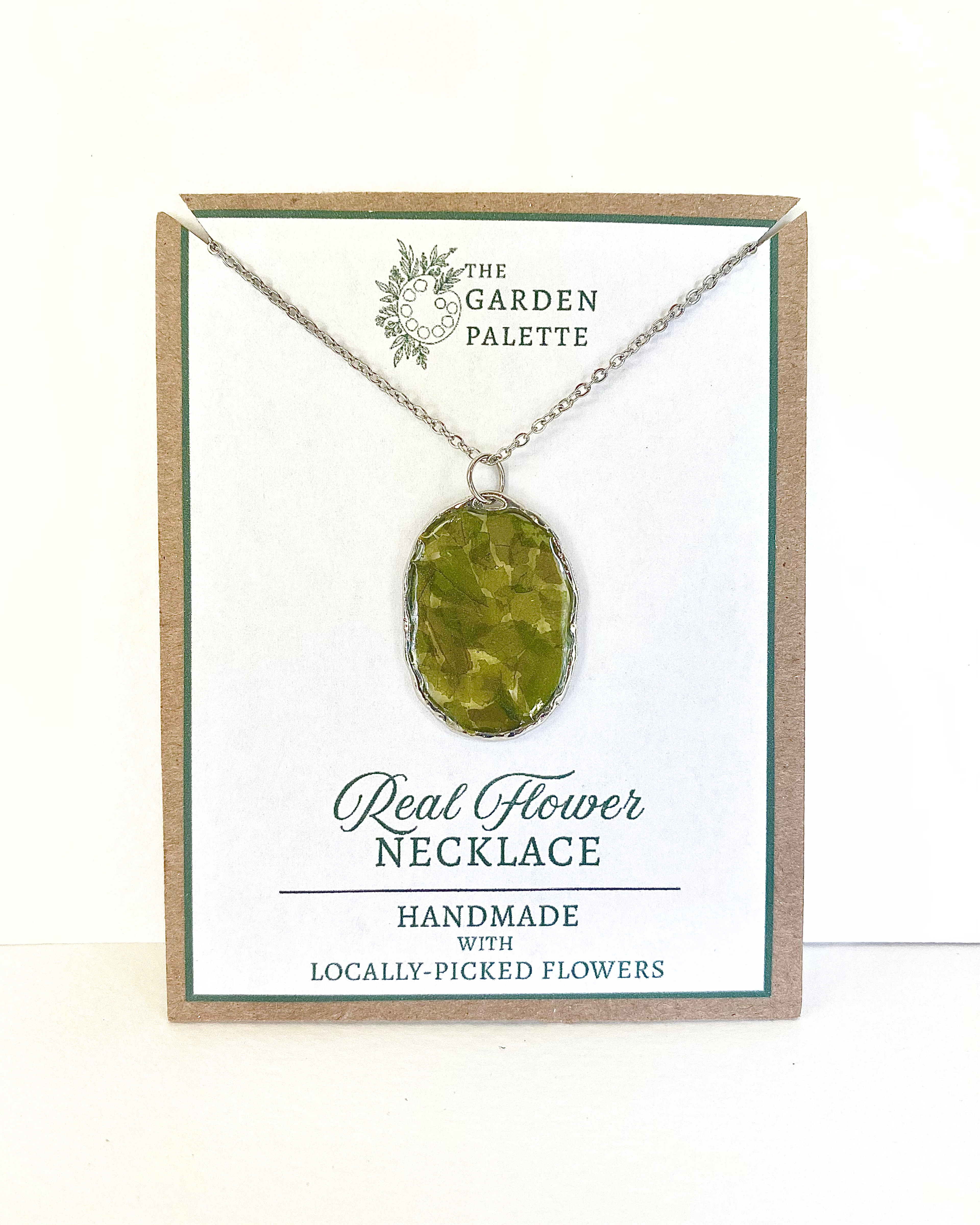 A silver-toned necklace chain with an oval bezel setting containing pressed green leave pieces, displayed on a branded card that reads 'The Garden Palette' and 'Real Flower Necklace'.