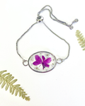 A silver adjustable chain bracelet featuring an oval pendant with a preserved purple flower and small white flowers, displayed on a white background with decorative green fern leaves.
