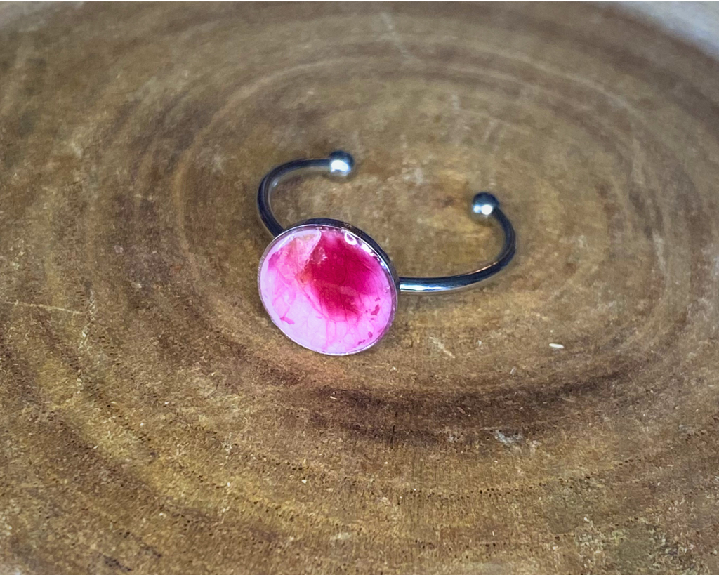 A silver-toned ring with a round bezel setting containing a pink petal, displayed on a rustic wood background.