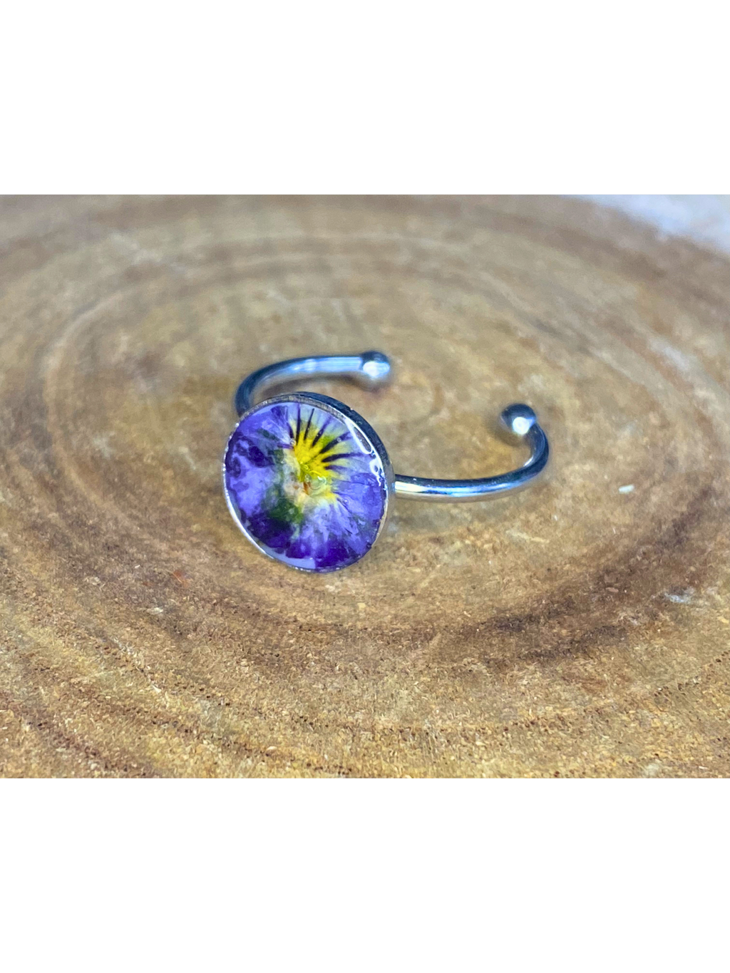 A silver-toned ring with a round bezel setting containing a blue and yellow flower, displayed on a rustic wood background