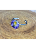 A silver-toned ring with a round bezel setting containing a blue and yellow flower, displayed on a rustic wood background