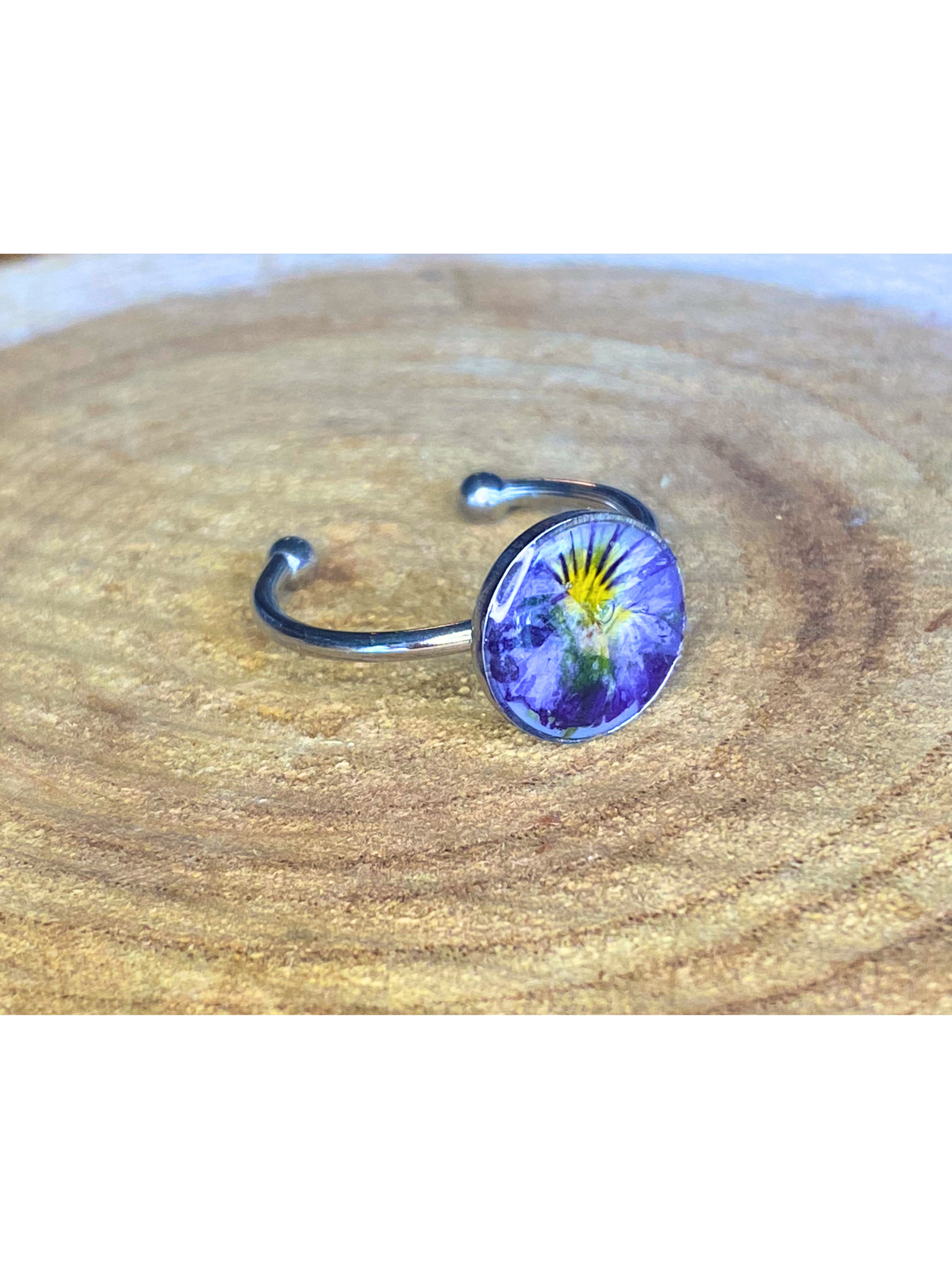 A silver-toned ring with a round bezel setting containing a blue and yellow flower, displayed on a rustic wood background