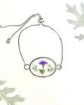 A silver chain bracelet with an oval pendant containing a small purple and white flowers under clear resin, displayed on a white background with decorative leaves.