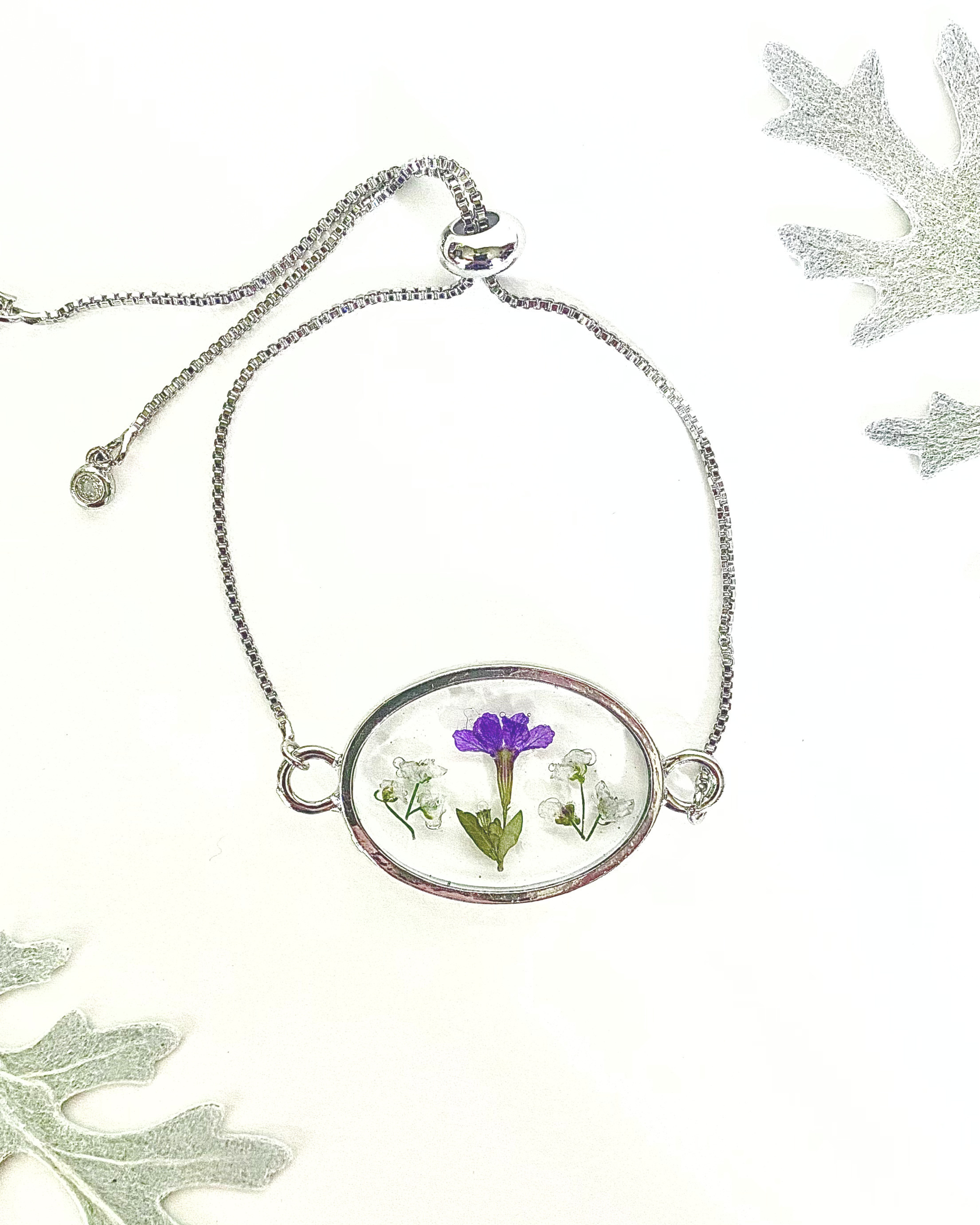 A silver chain bracelet with an oval pendant containing a small purple and white flowers under clear resin, displayed on a white background with decorative leaves.